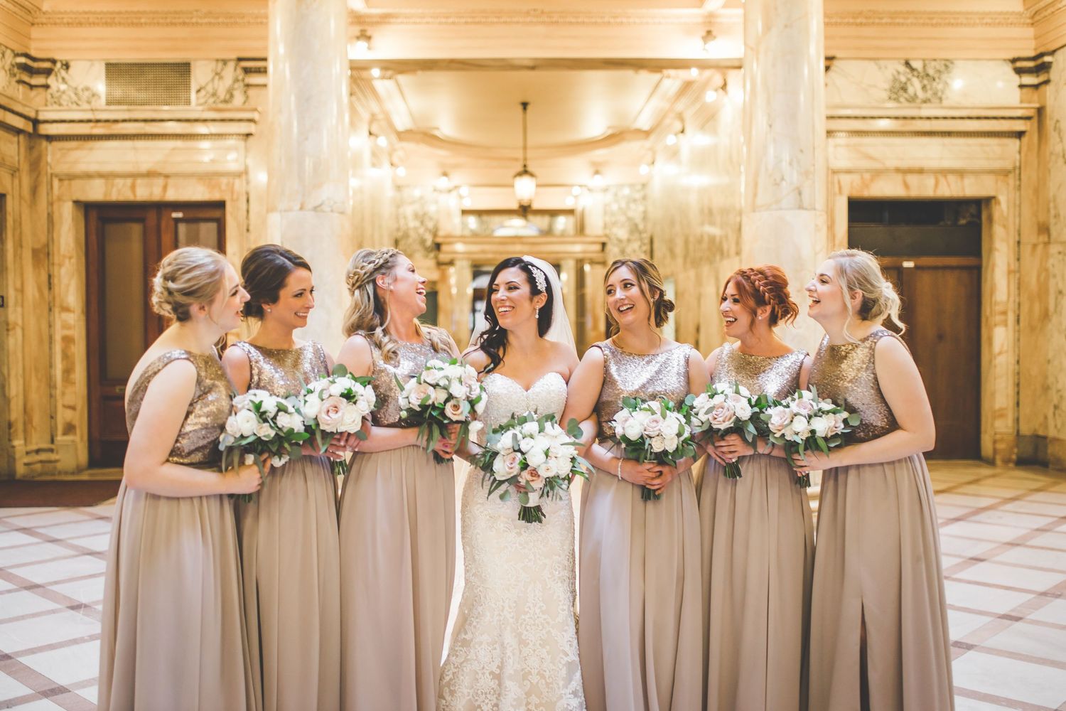 Bridesmaids in champagne colored dresses hold white floral bouquets in an elegant indoor venue.