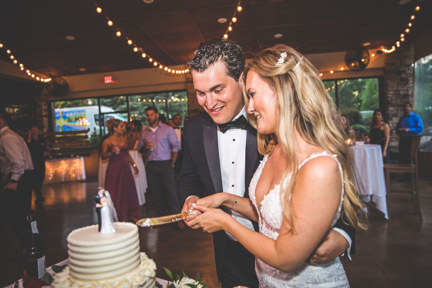 A couple cuts their white wedding cake together under string lights at their reception.