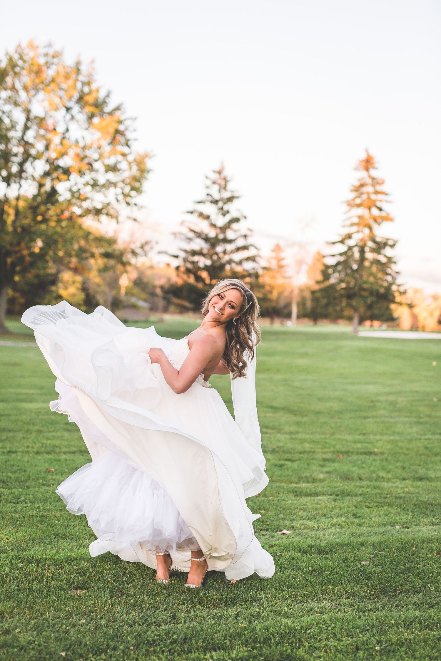 Bride in flowing white dress twirling gracefully on green lawn at sunset.
