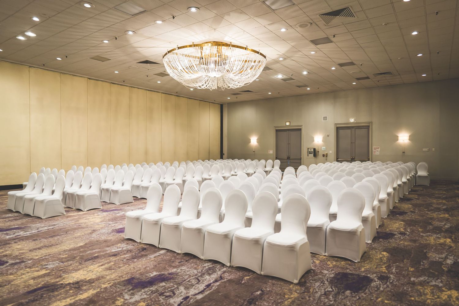 Large wedding venue ballroom with rows of white chairs and elegant crystal chandeliers.