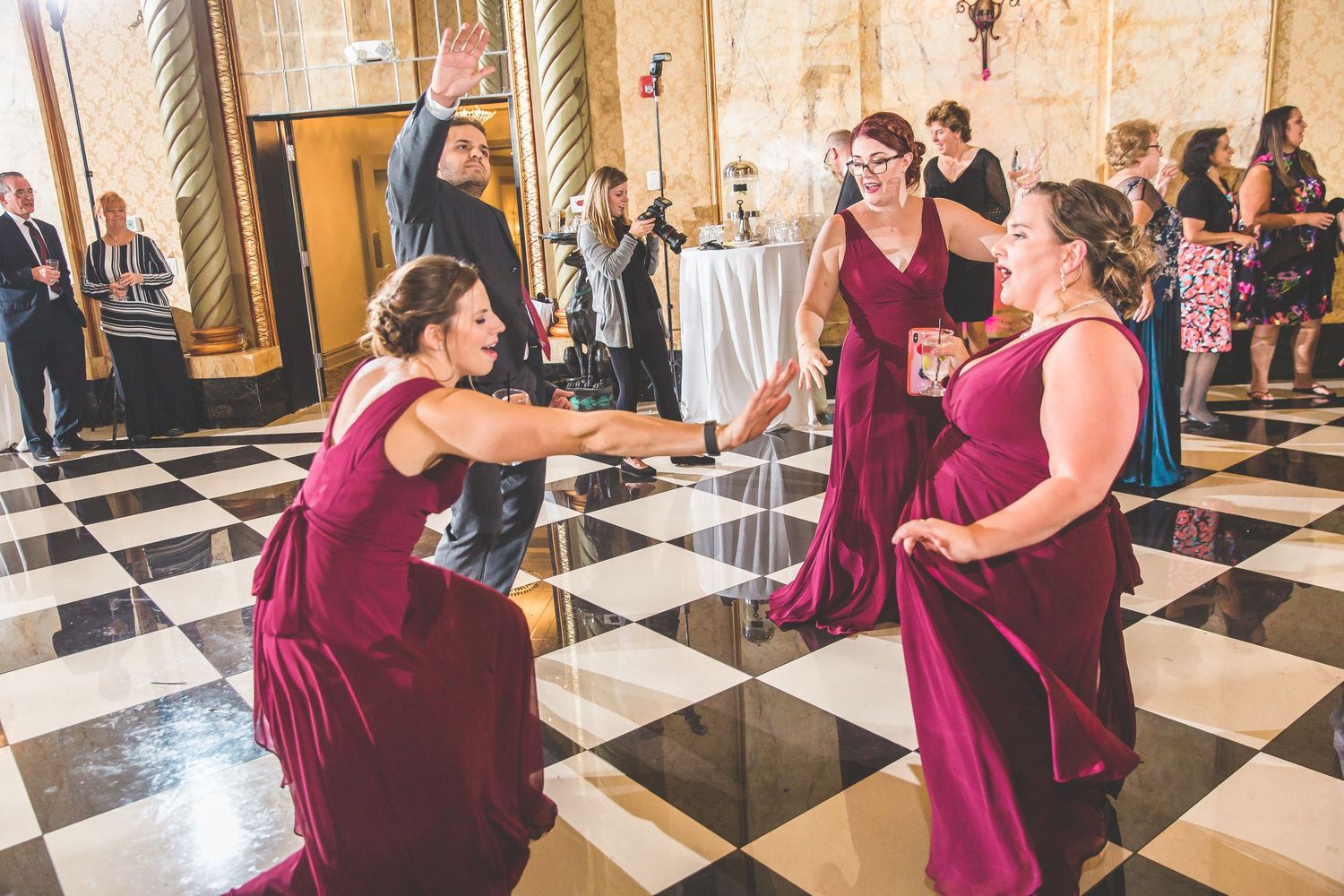Bridesmaids in burgundy dresses dancing together on a black and white checkered floor.