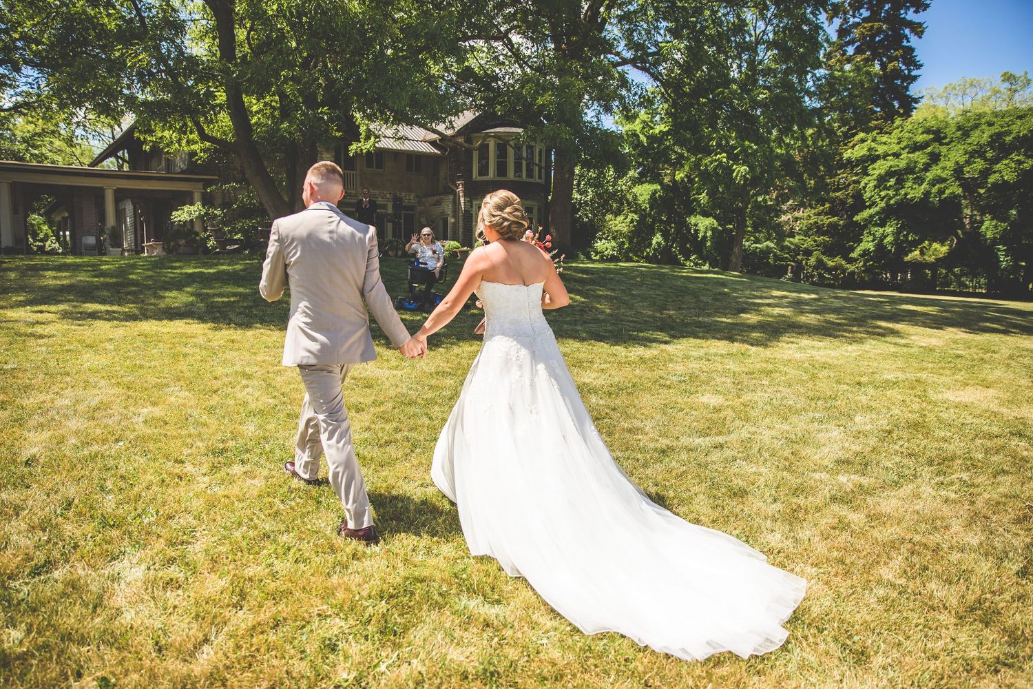 A couple in wedding attire walks hand in hand across a sunny garden lawn.