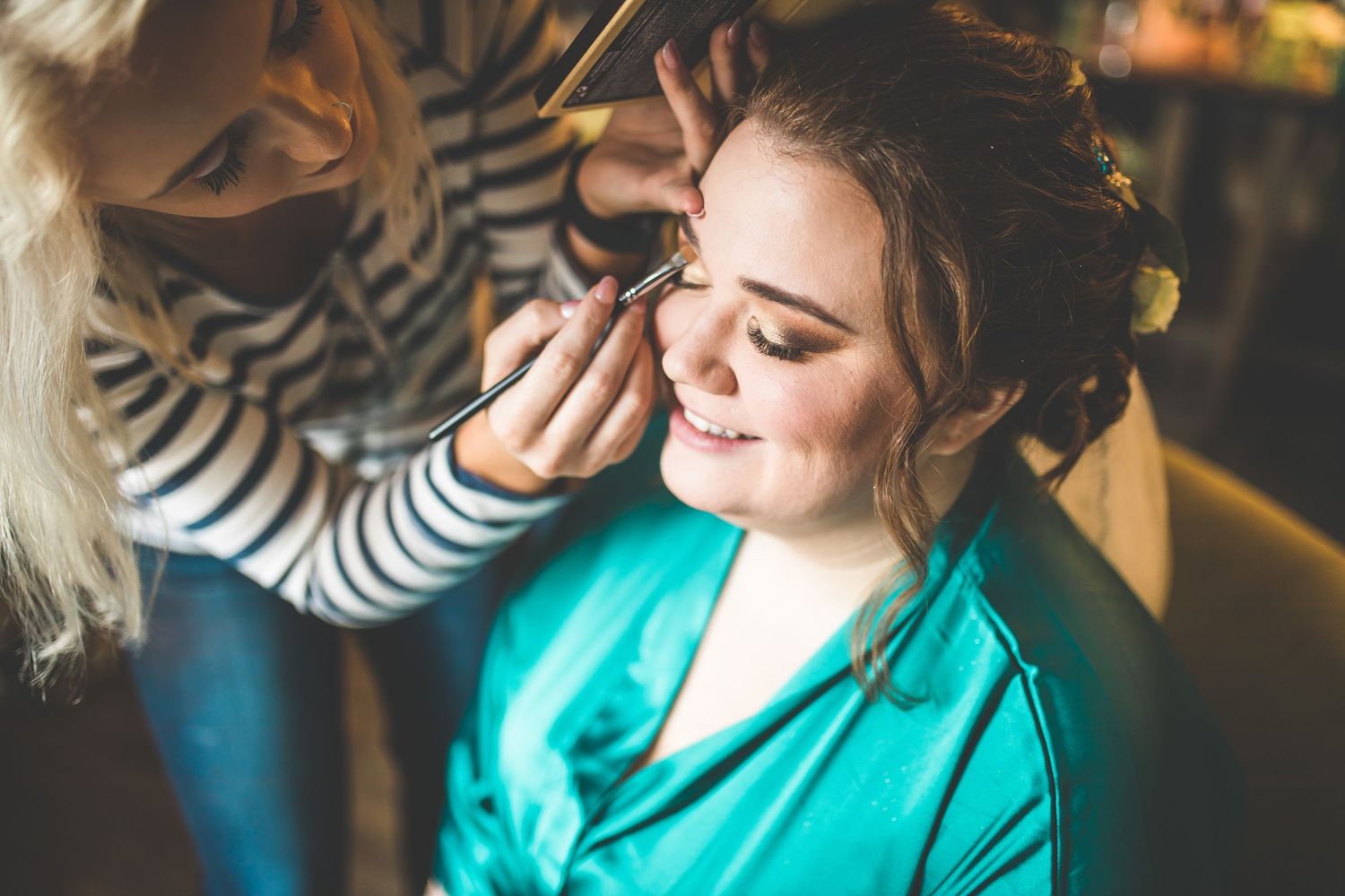 A woman in turquoise getting her makeup professionally applied during wedding preparations.