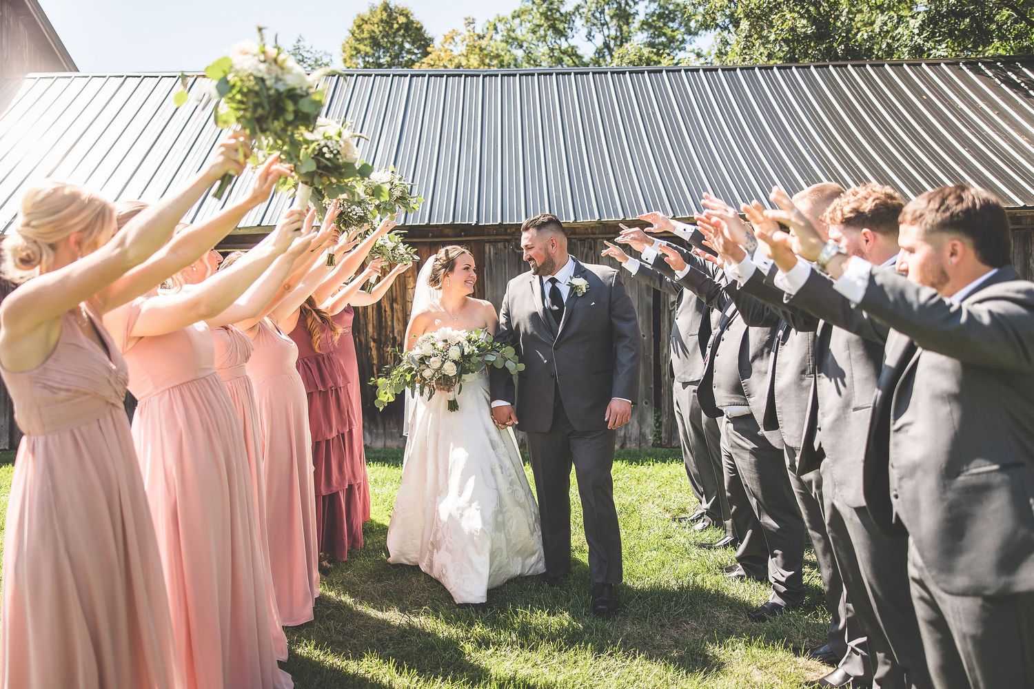 Wedding party forms two lines with bridesmaids in pink and groomsmen in gray celebrating the newlyweds outdoors.