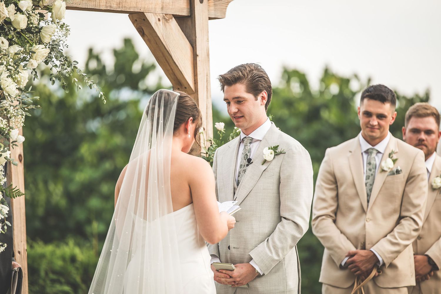 Couple exchanges vows under wooden arbor during outdoor wedding ceremony.