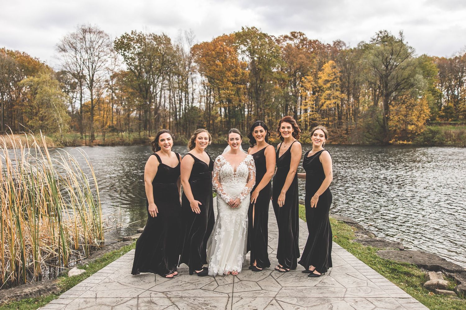 Wedding party in black dresses poses with bride on stone path near autumn lake.