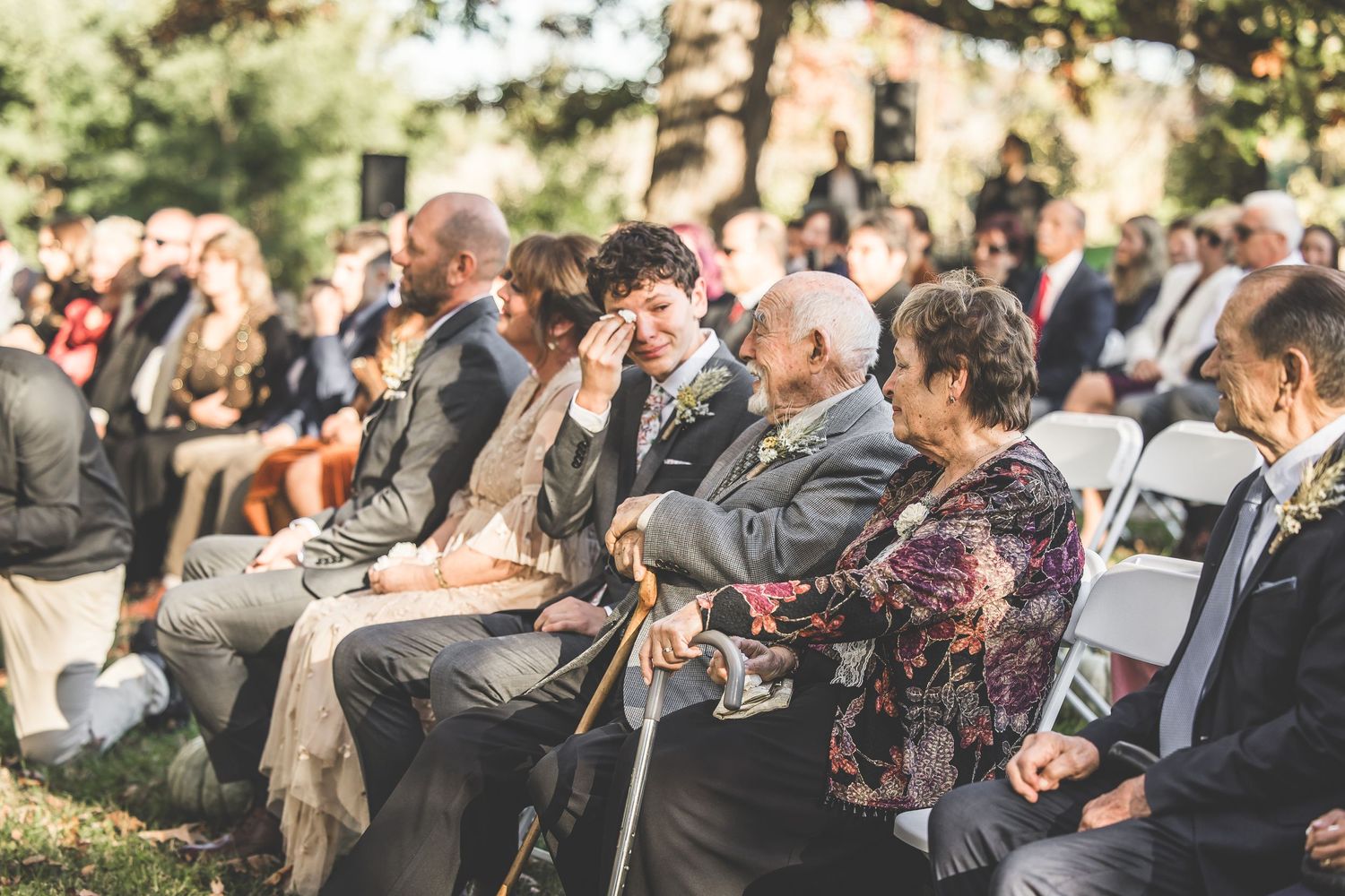 Wedding guests seated outdoors at a fall ceremony with golden sunlight filtering through trees.