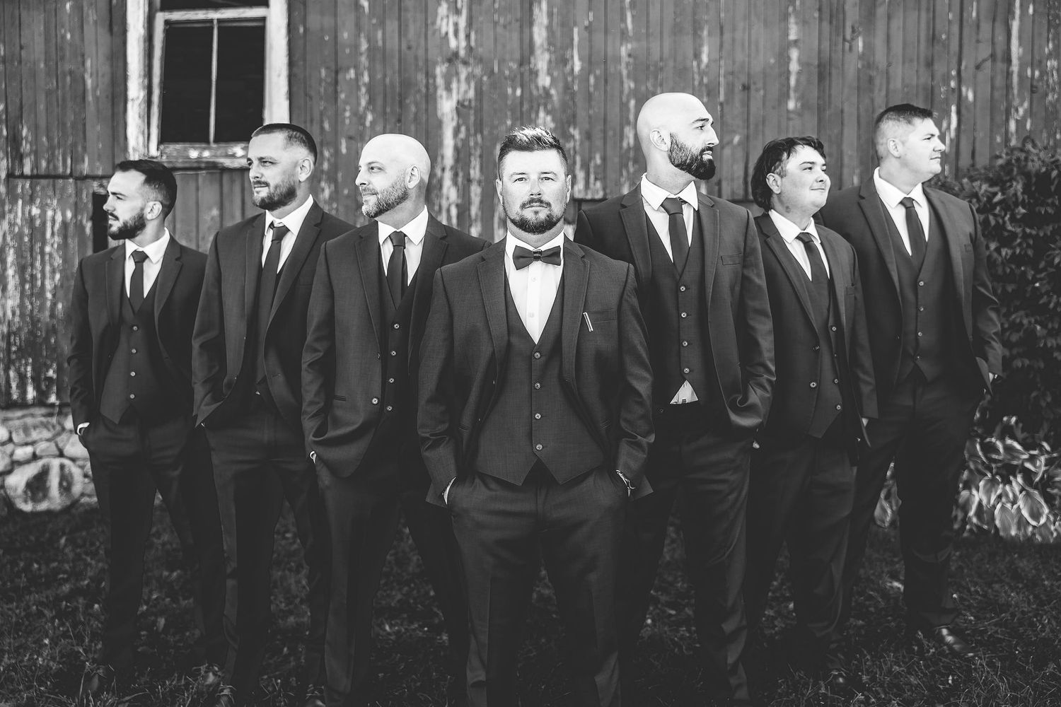 Black and white portrait of groomsmen lined up along vintage barn wall.