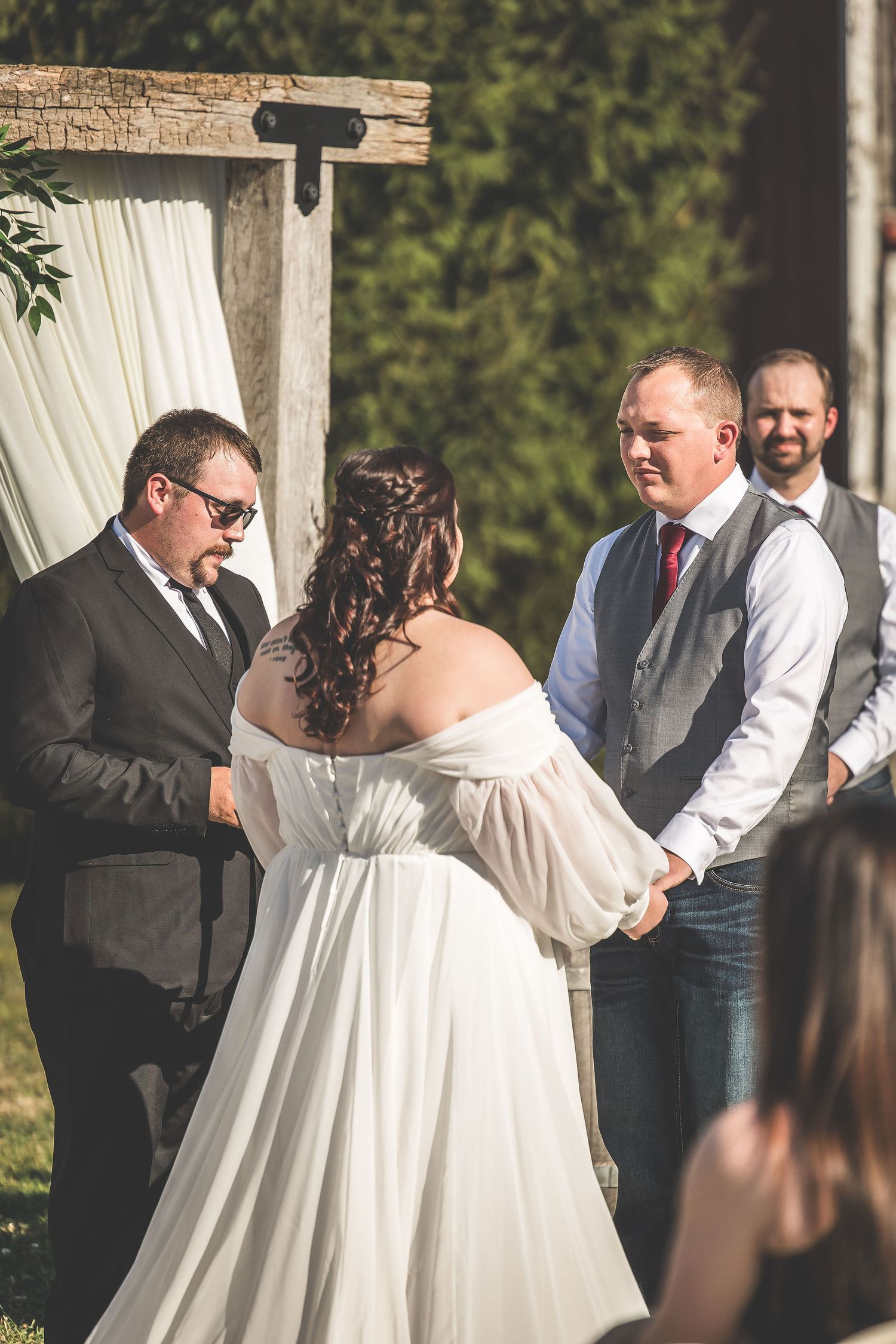 Wedding ceremony taking place under an outdoor pavilion with white drapes and autumn cornfield in the background.