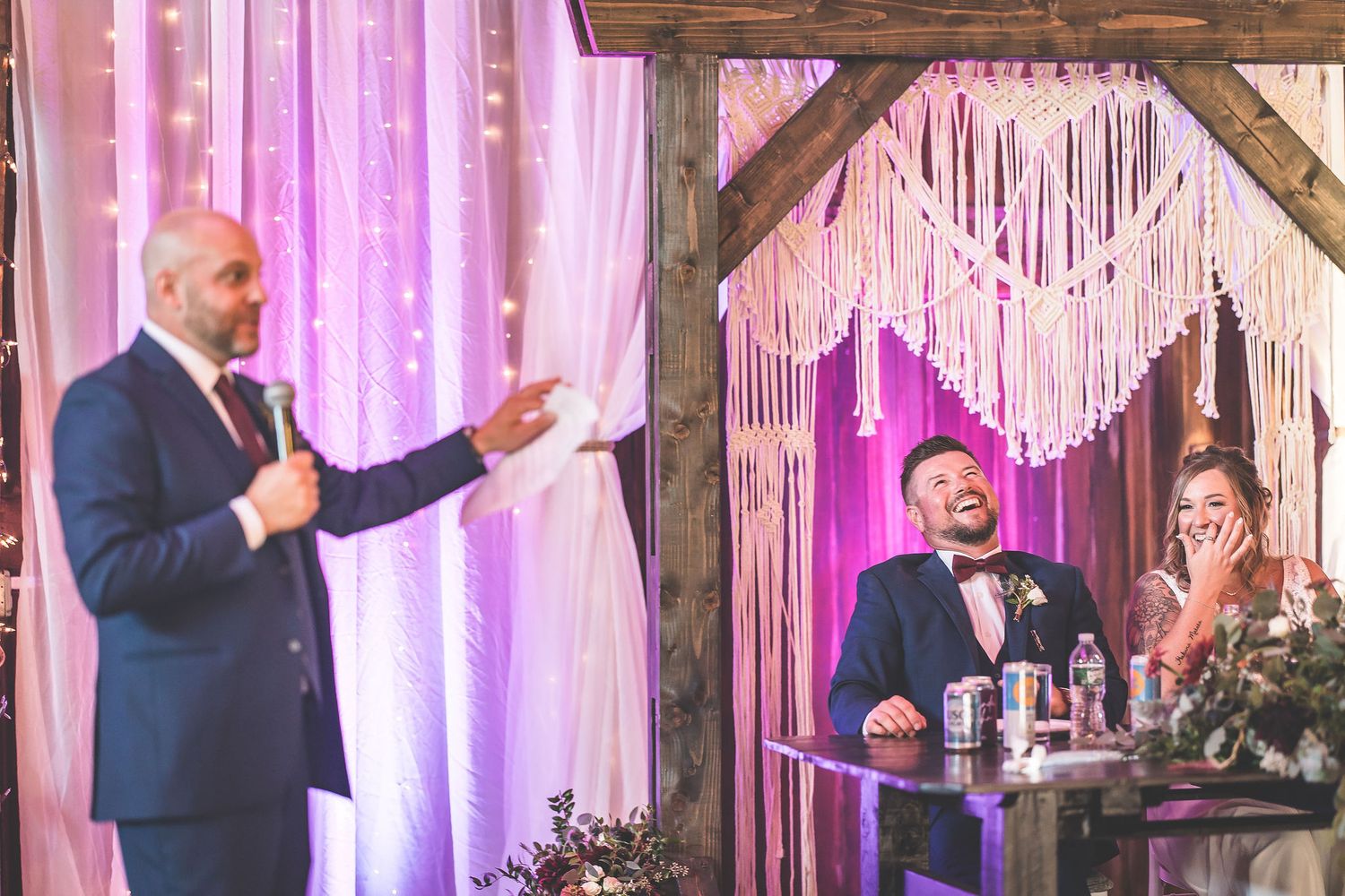 Wedding reception speeches under rustic wooden beams with macrame backdrop and purple lighting.