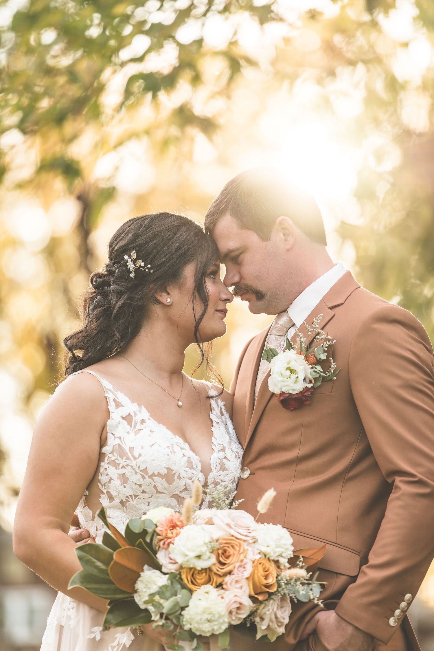 Romantic wedding couple embraces in golden sunlight while holding a bouquet of blush and white flowers.