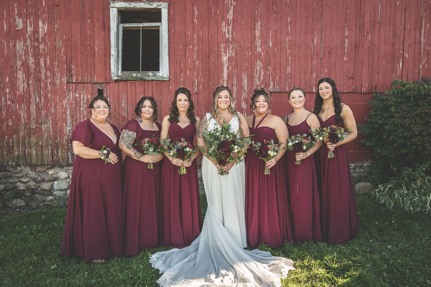 Wedding party in burgundy dresses poses in front of rustic red barn.