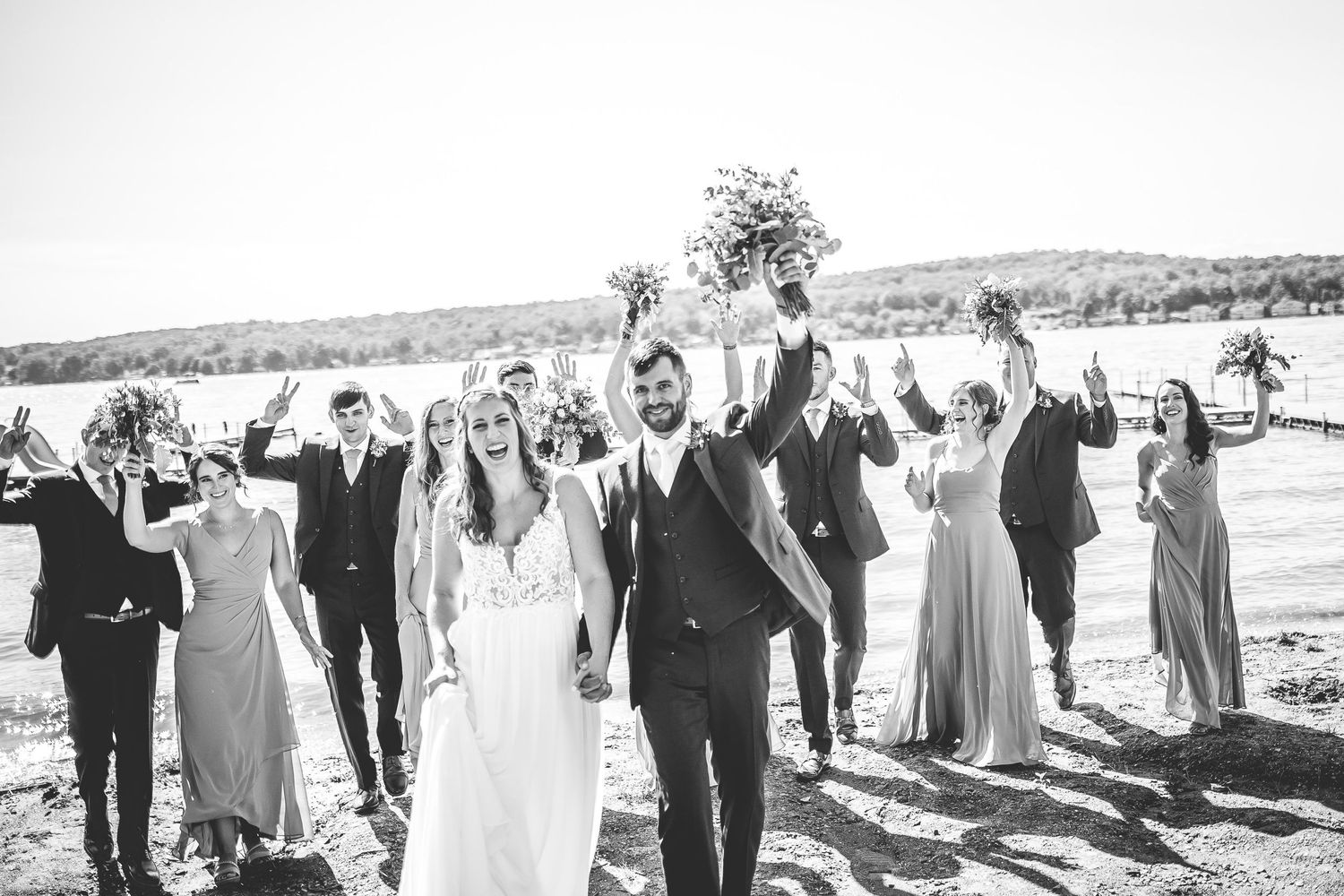 Wedding party celebrates joyfully outdoors with raised arms against a scenic landscape background.