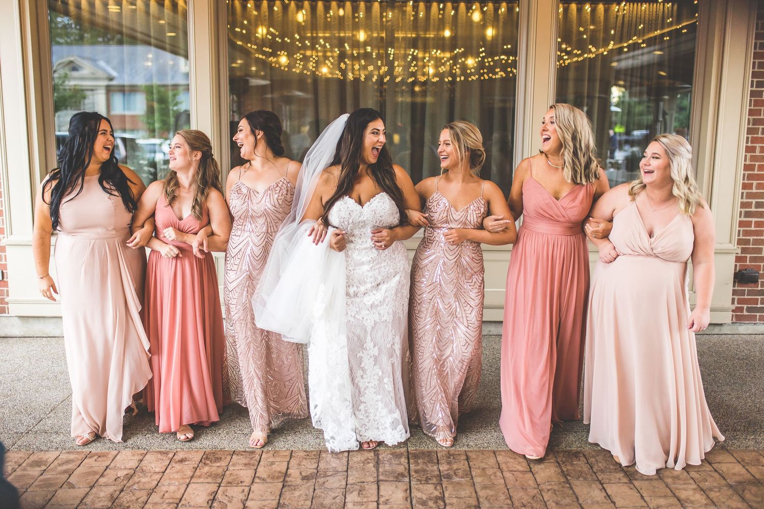 Bridal party in various shades of pink dresses poses together under string lights.