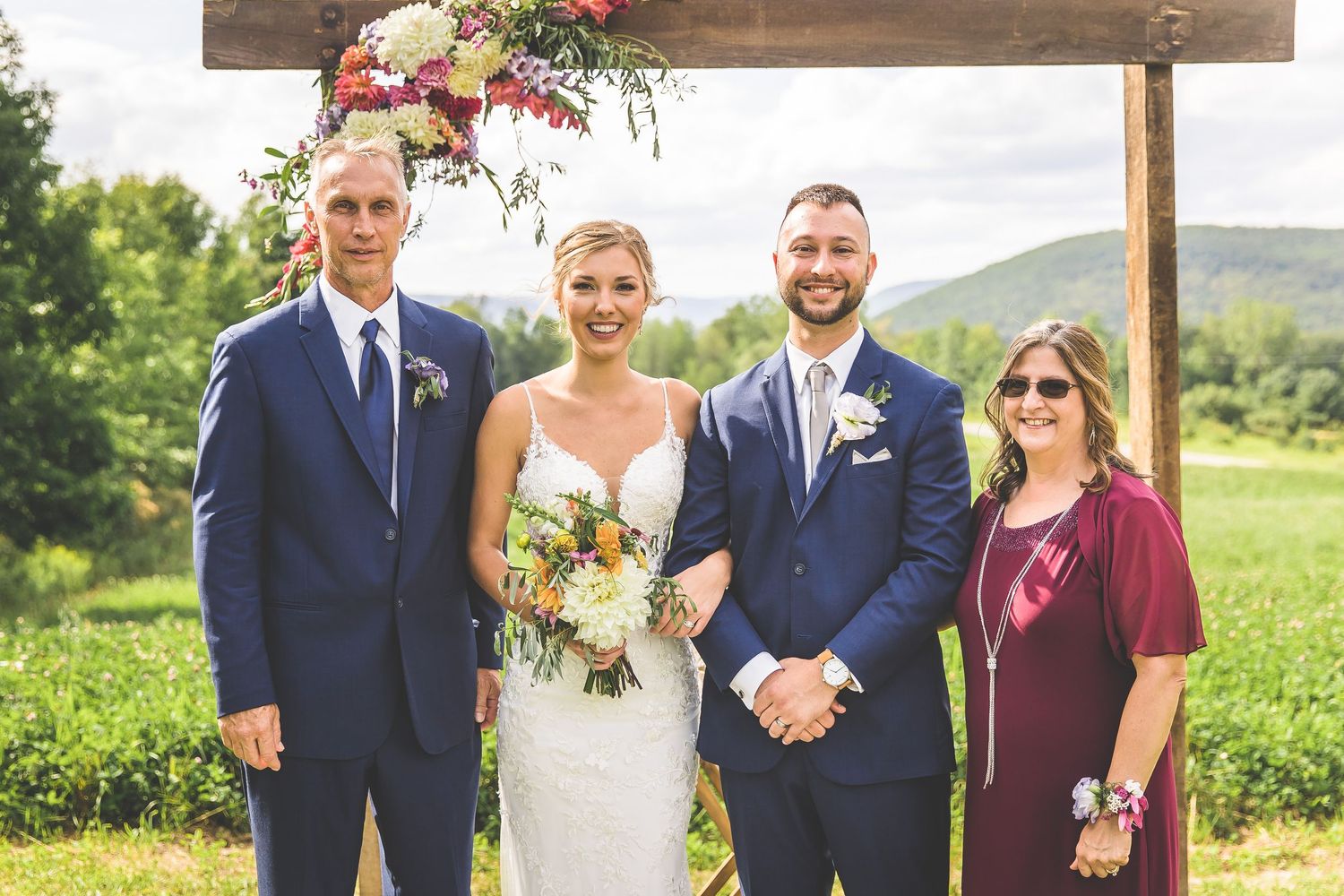 Wedding party members in navy and burgundy attire pose together under wooden ceremony arch.