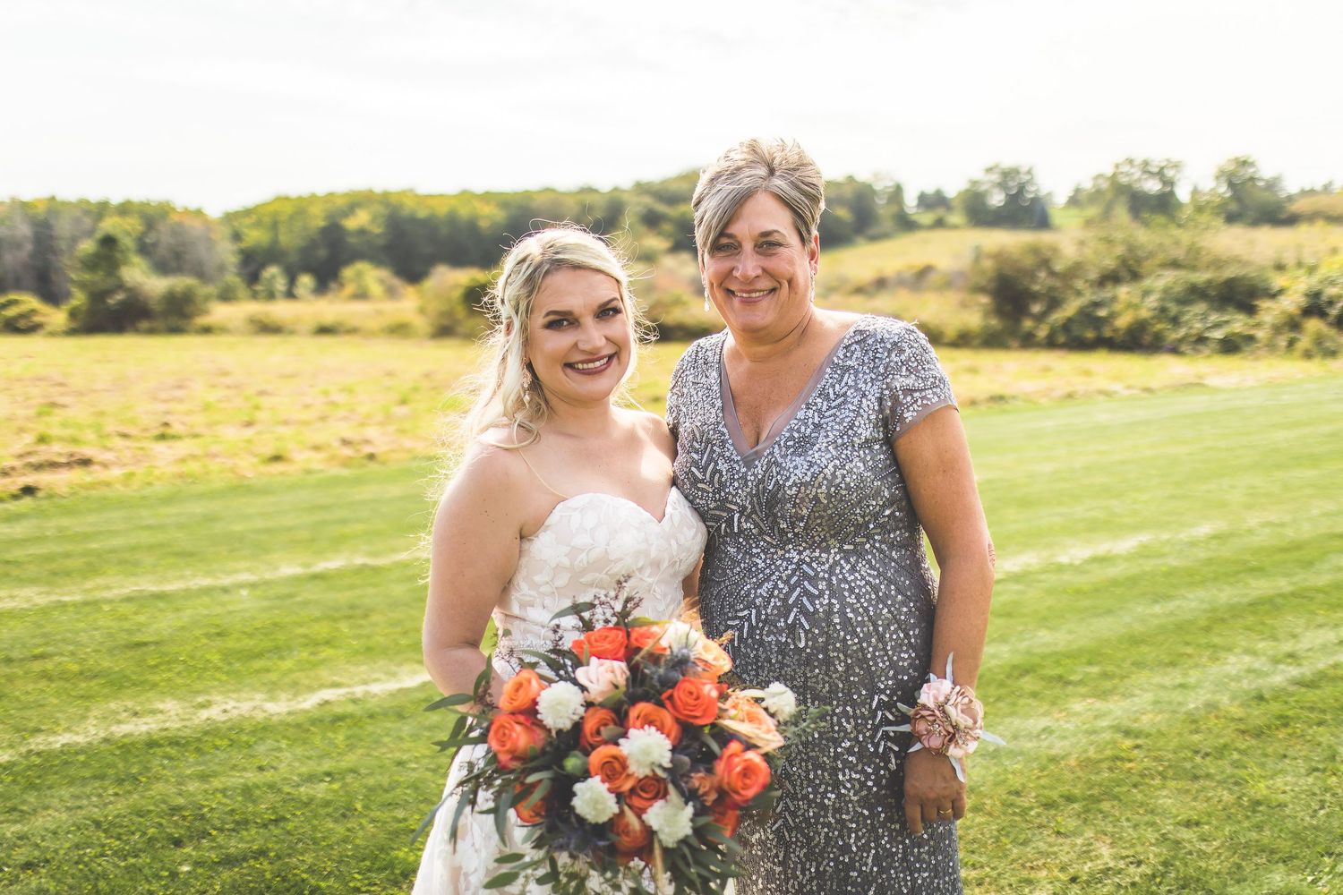 Two women in formal attire hold vibrant coral and peach bouquets in an outdoor setting.