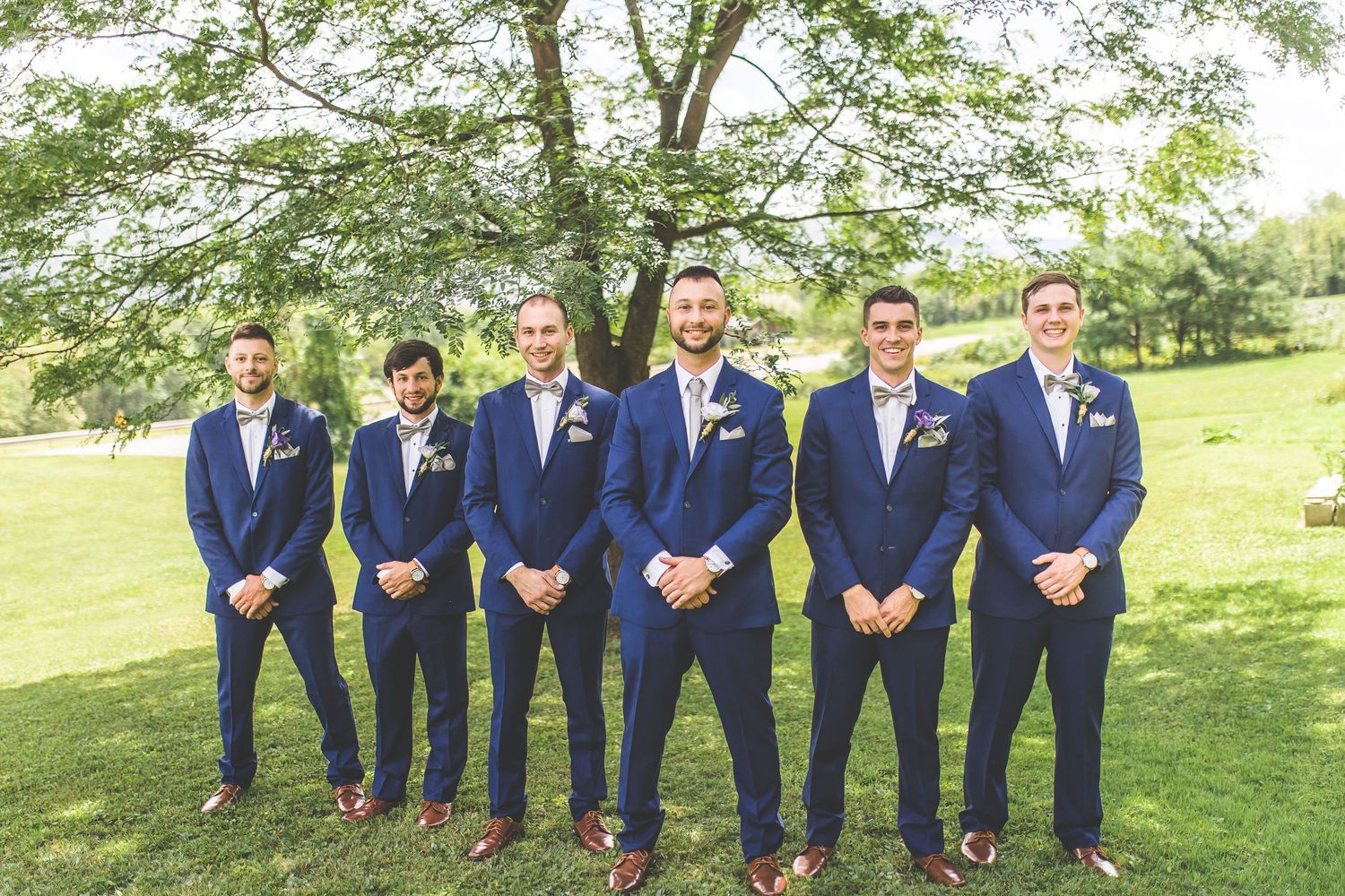 Wedding party in navy suits poses under green trees on manicured lawn.