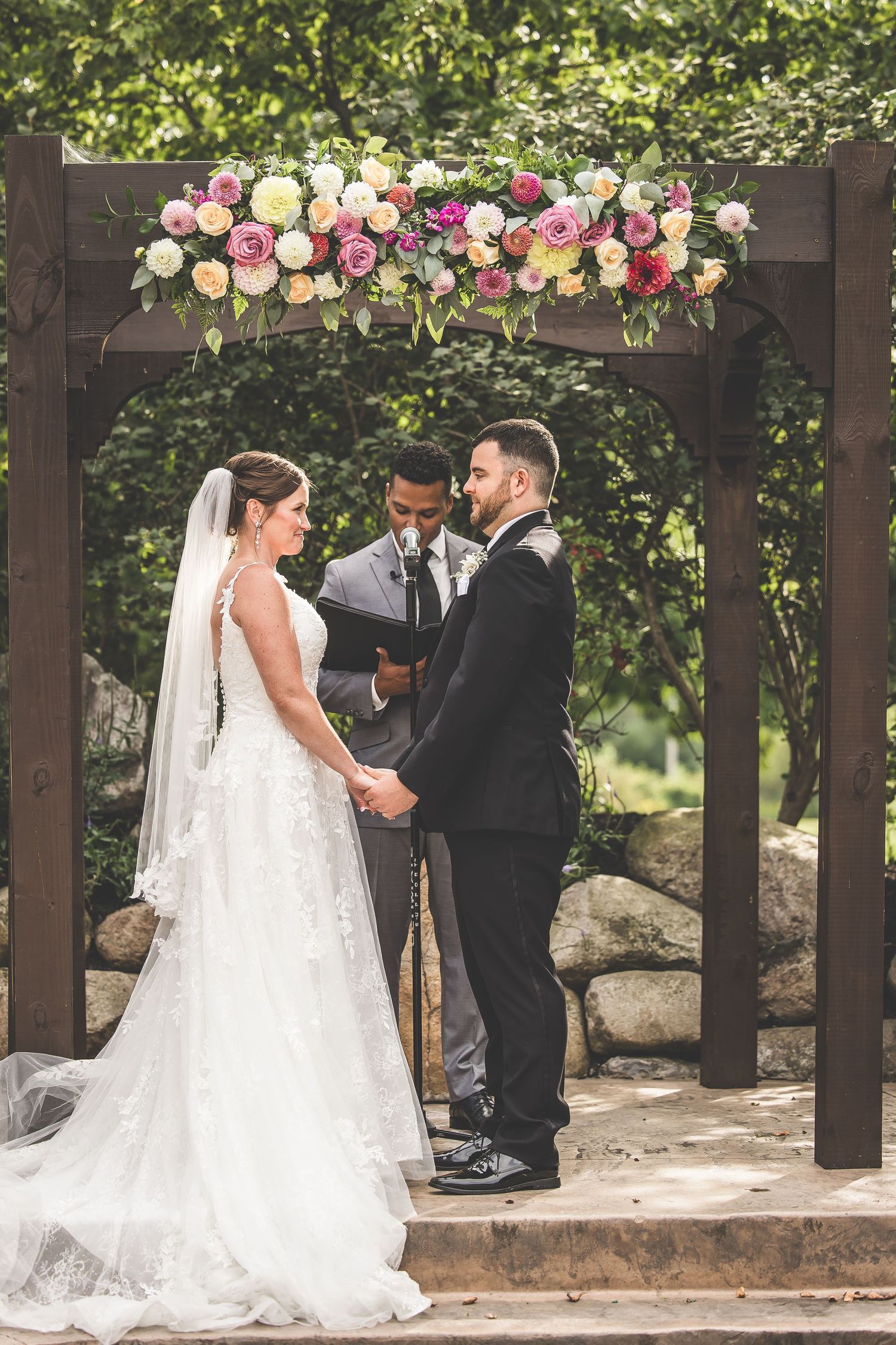 Wedding ceremony under a wooden pergola decorated with pink and white floral arrangements.