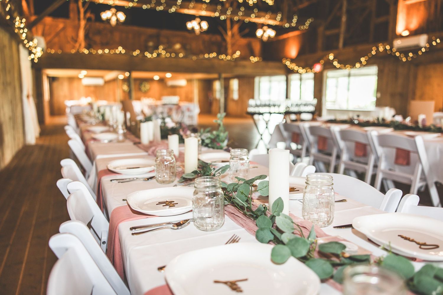 Long banquet tables set with white linens and place settings in a rustic wooden barn venue.