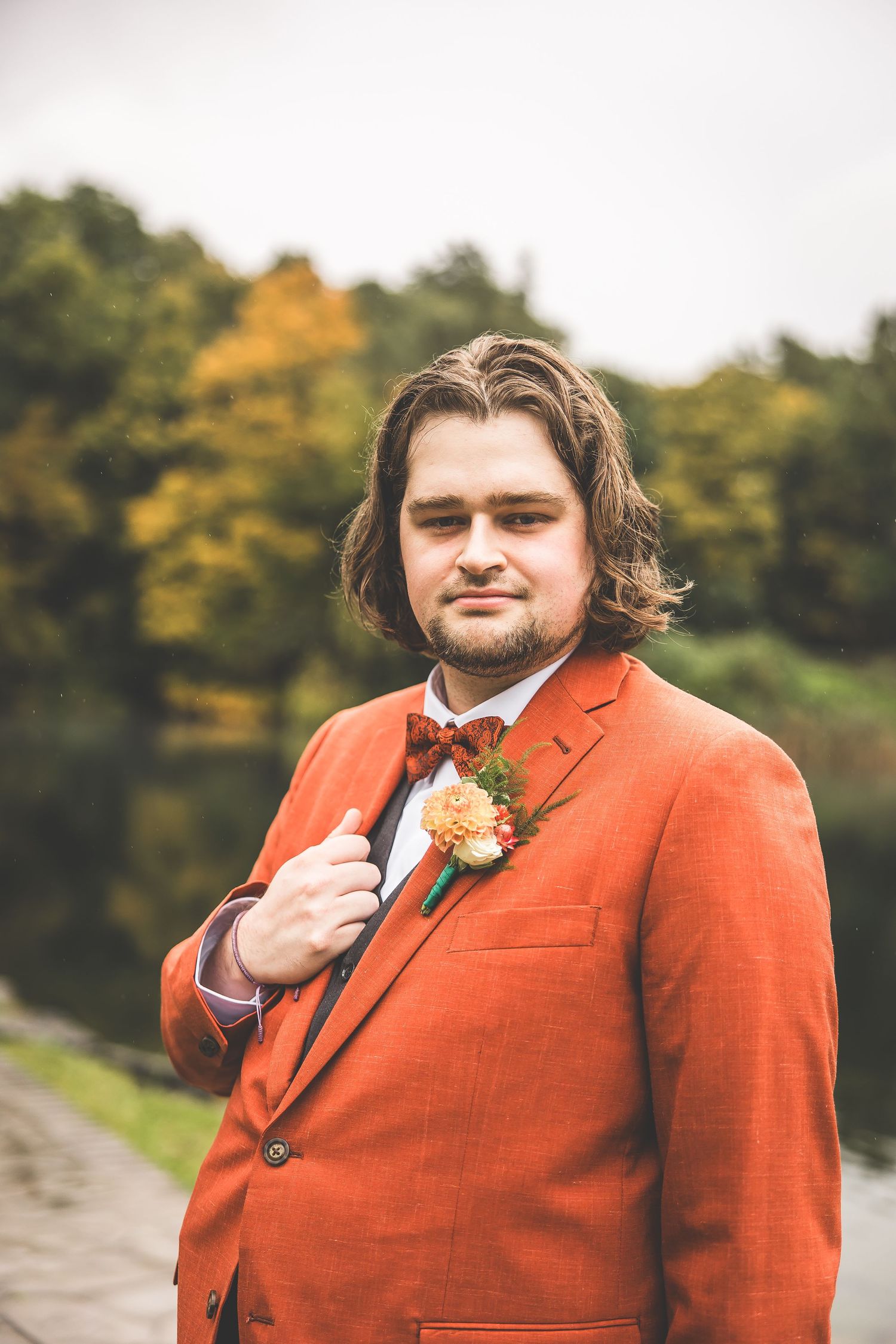 A groom in a bright orange suit with bow tie poses outdoors during autumn.