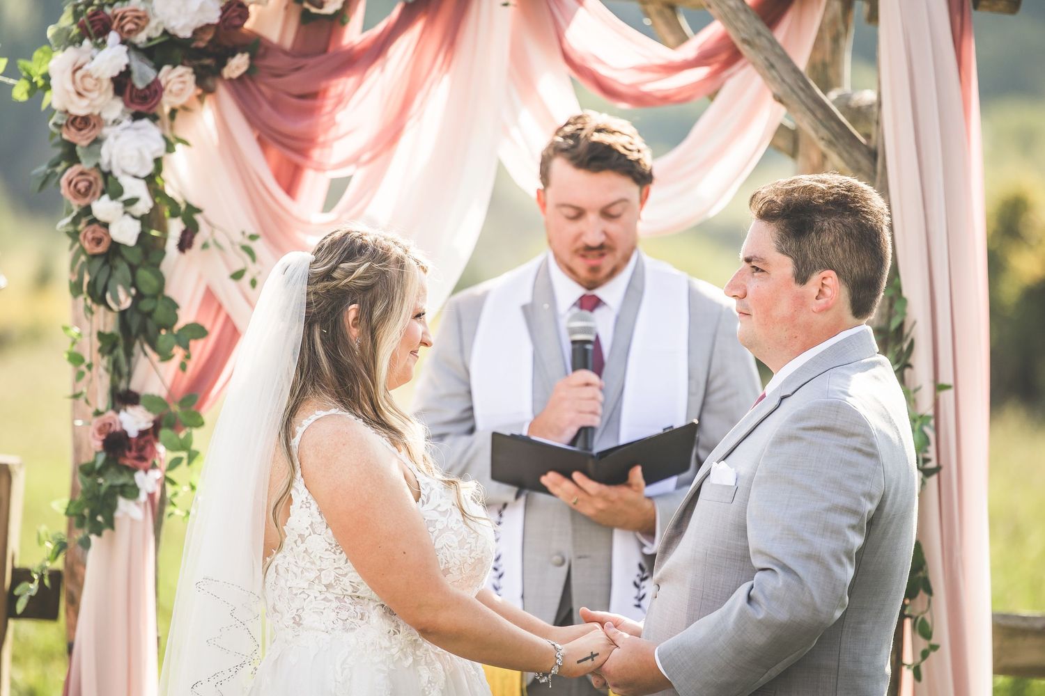 Wedding couple exchanges vows beneath a rustic wooden arch decorated with pink and white flowers.
