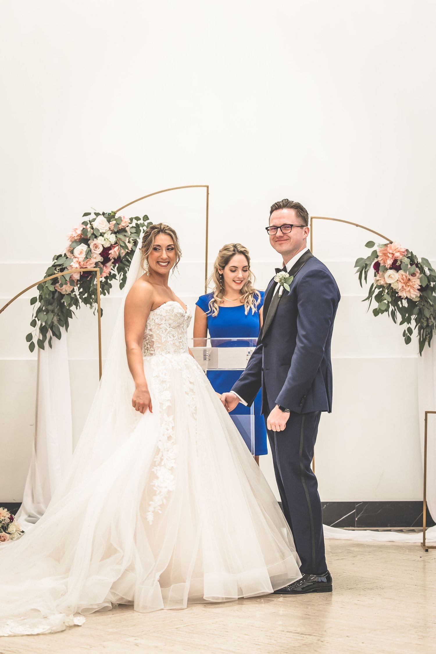 Wedding couple posing under copper arch with floral arrangements in white venue.