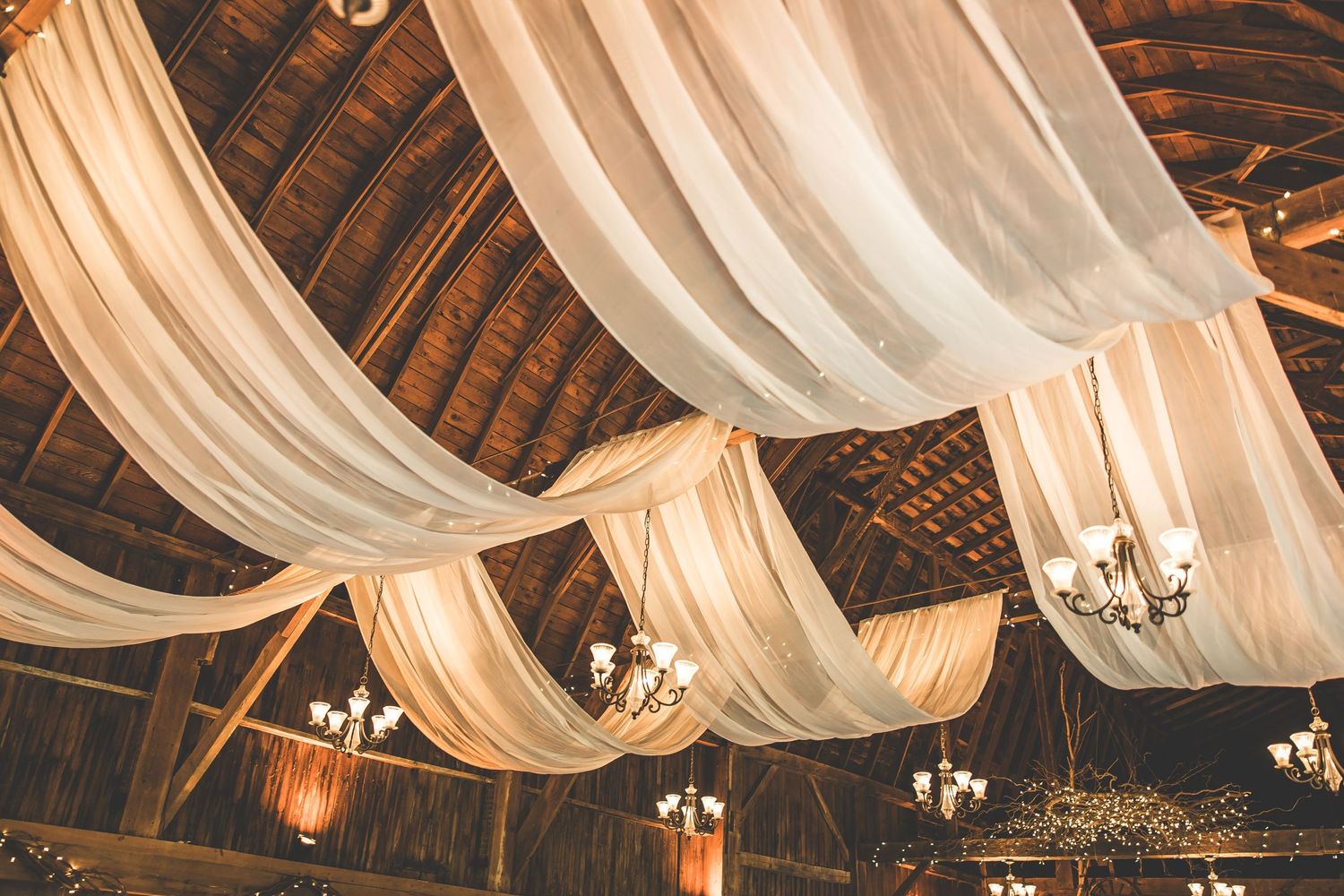 Romantic white fabric draping adorns wooden beams with crystal chandeliers in a rustic reception venue.
