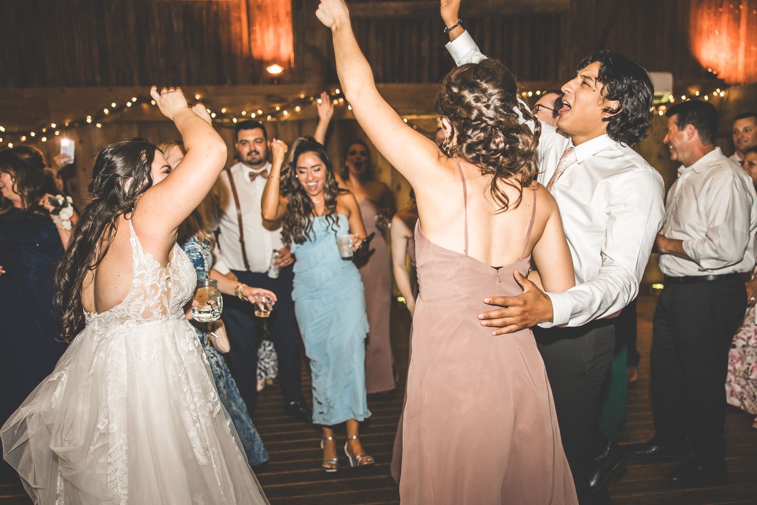 Wedding party dancing in a rustic venue illuminated by warm string lights and ambient lighting.