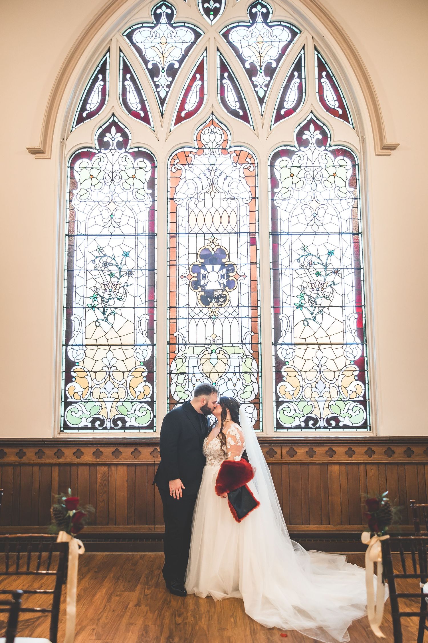 Wedding couple poses in front of ornate stained glass windows inside a historic church.