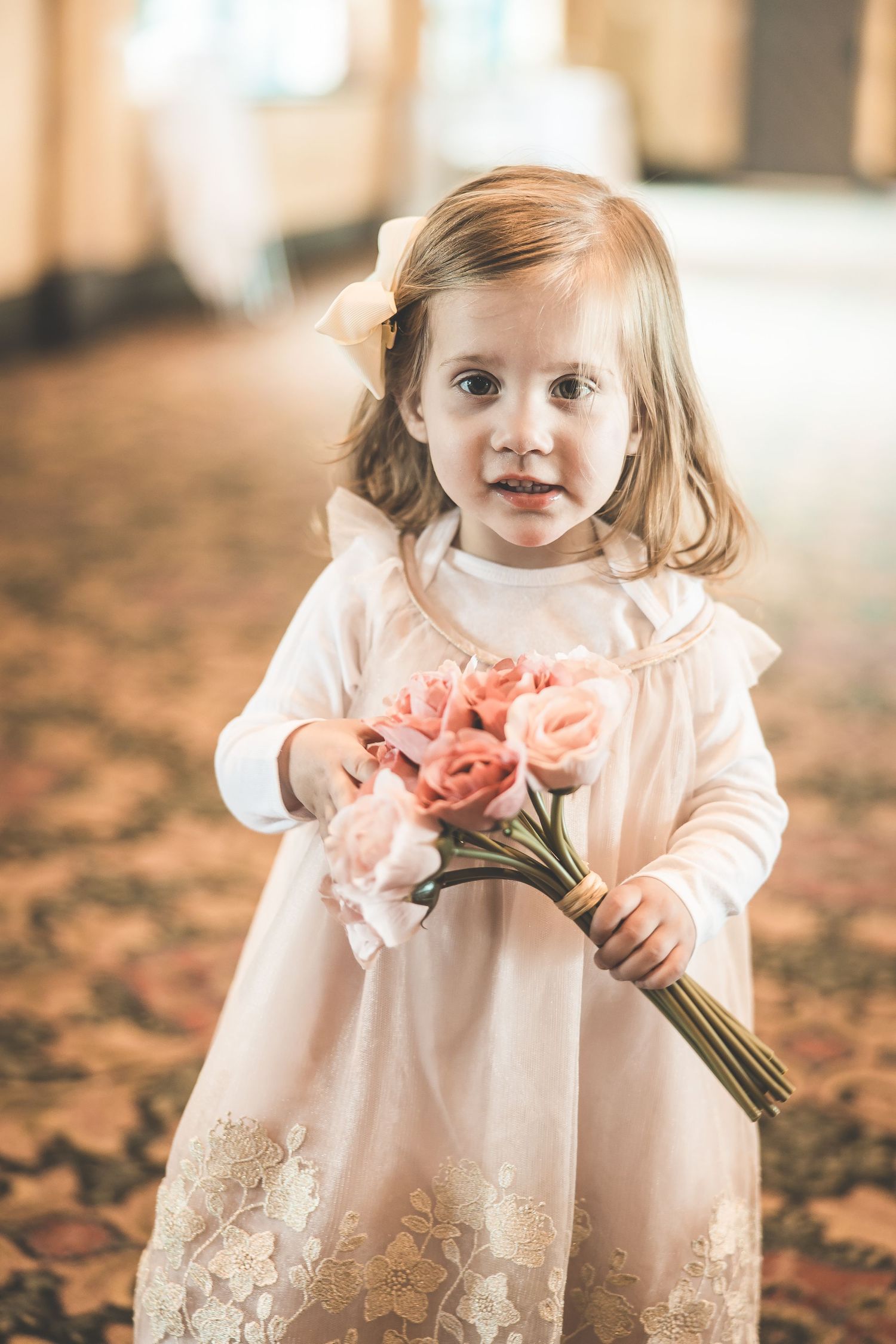Little flower girl in lace dress holds pink flowers at wedding.