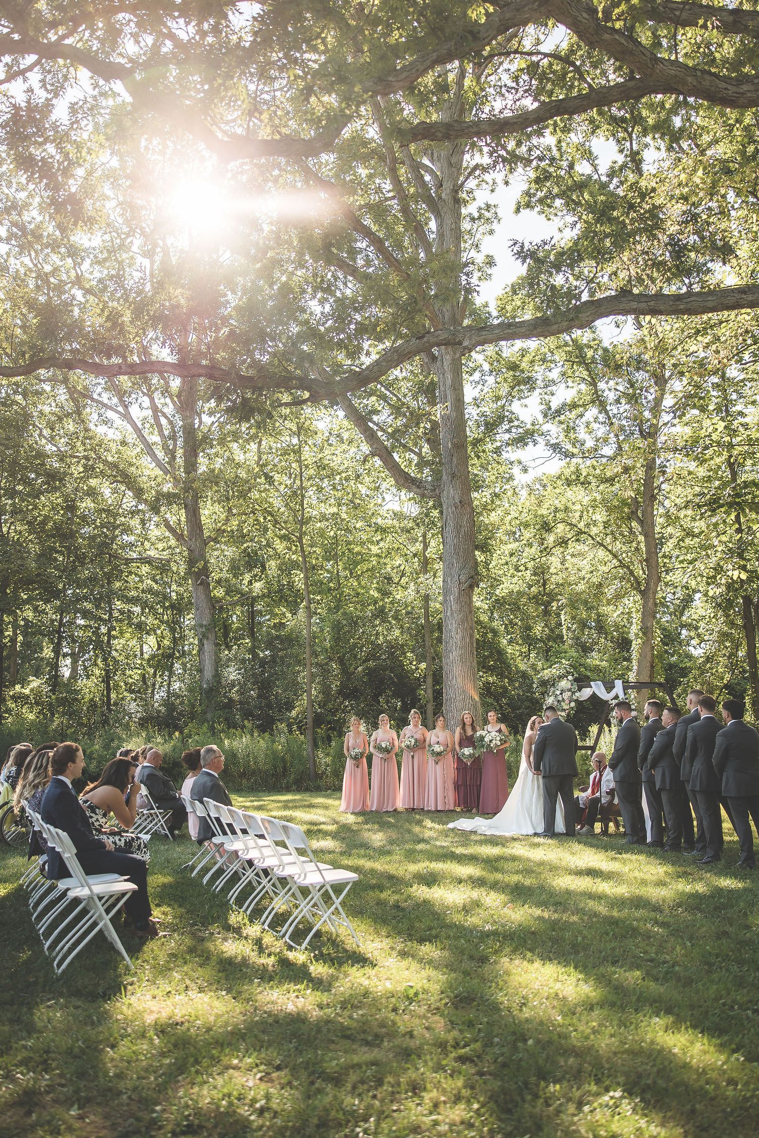 An outdoor wedding ceremony in progress under tall trees with sunlight streaming through the branches.
