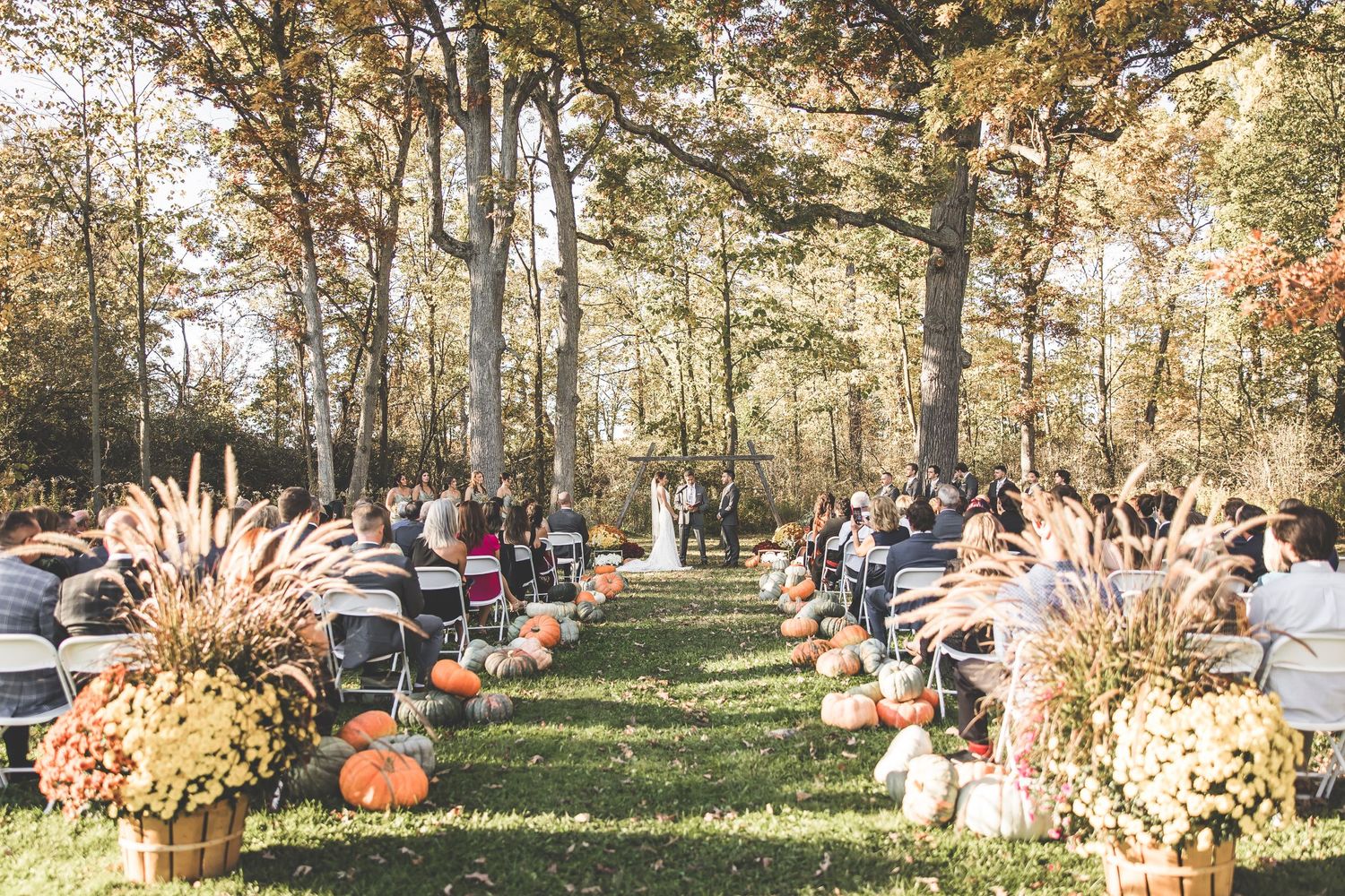 Outdoor fall wedding ceremony setup with pumpkins and dried grass decorations lining the aisle.