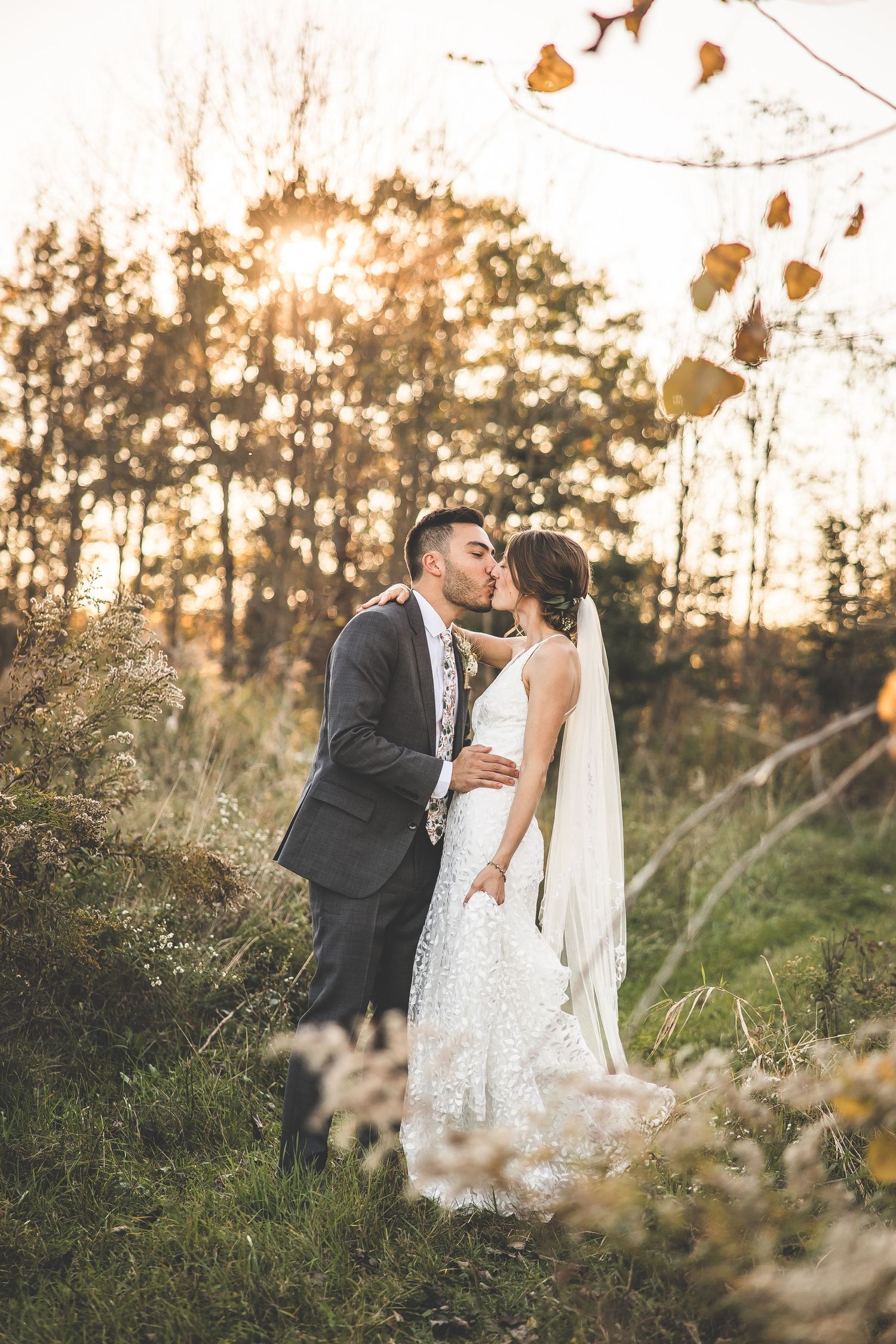 A romantic wedding couple embraces during golden hour sunset in a field with tall grasses and trees.