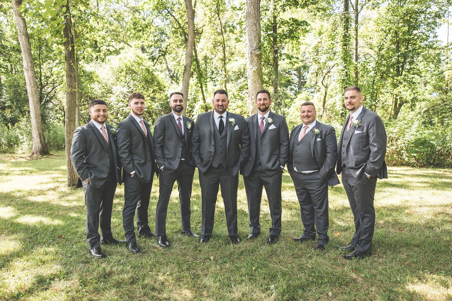 Wedding party groomsmen in grey suits stand in line against a natural forest backdrop.