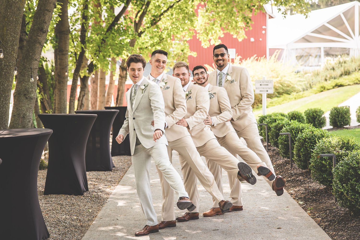 Groomsmen in light colored suits posing together under trees near a red building.