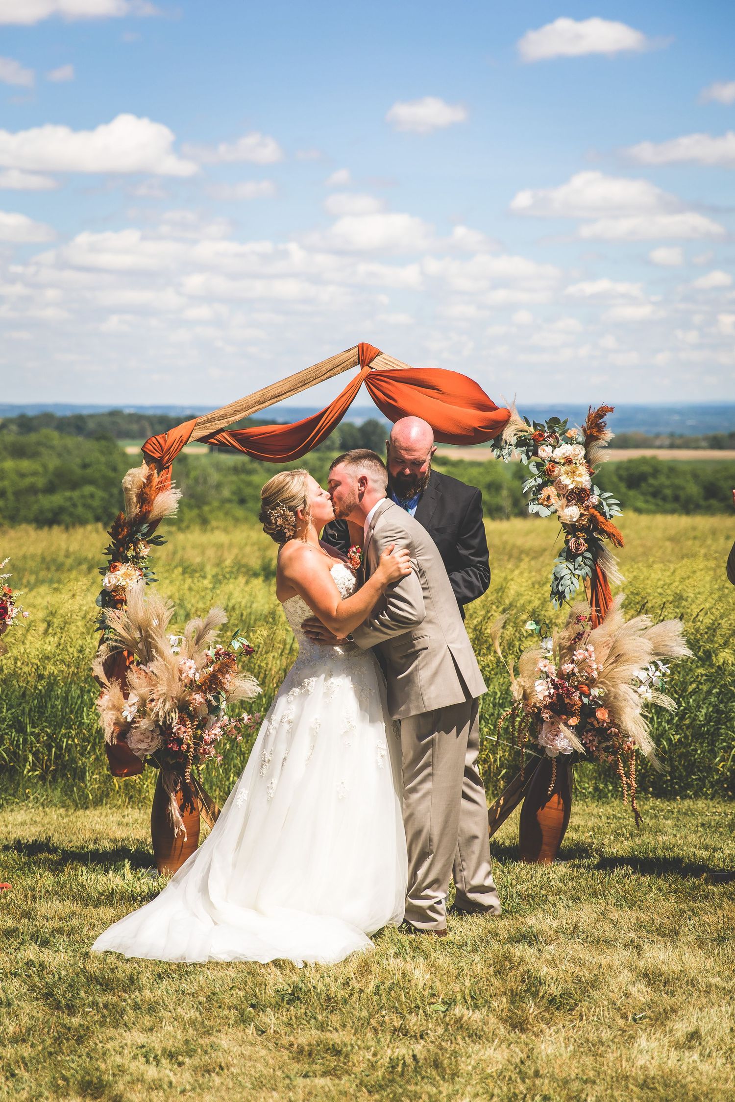 Outdoor hilltop wedding ceremony with orange fabric arch and natural landscape backdrop.