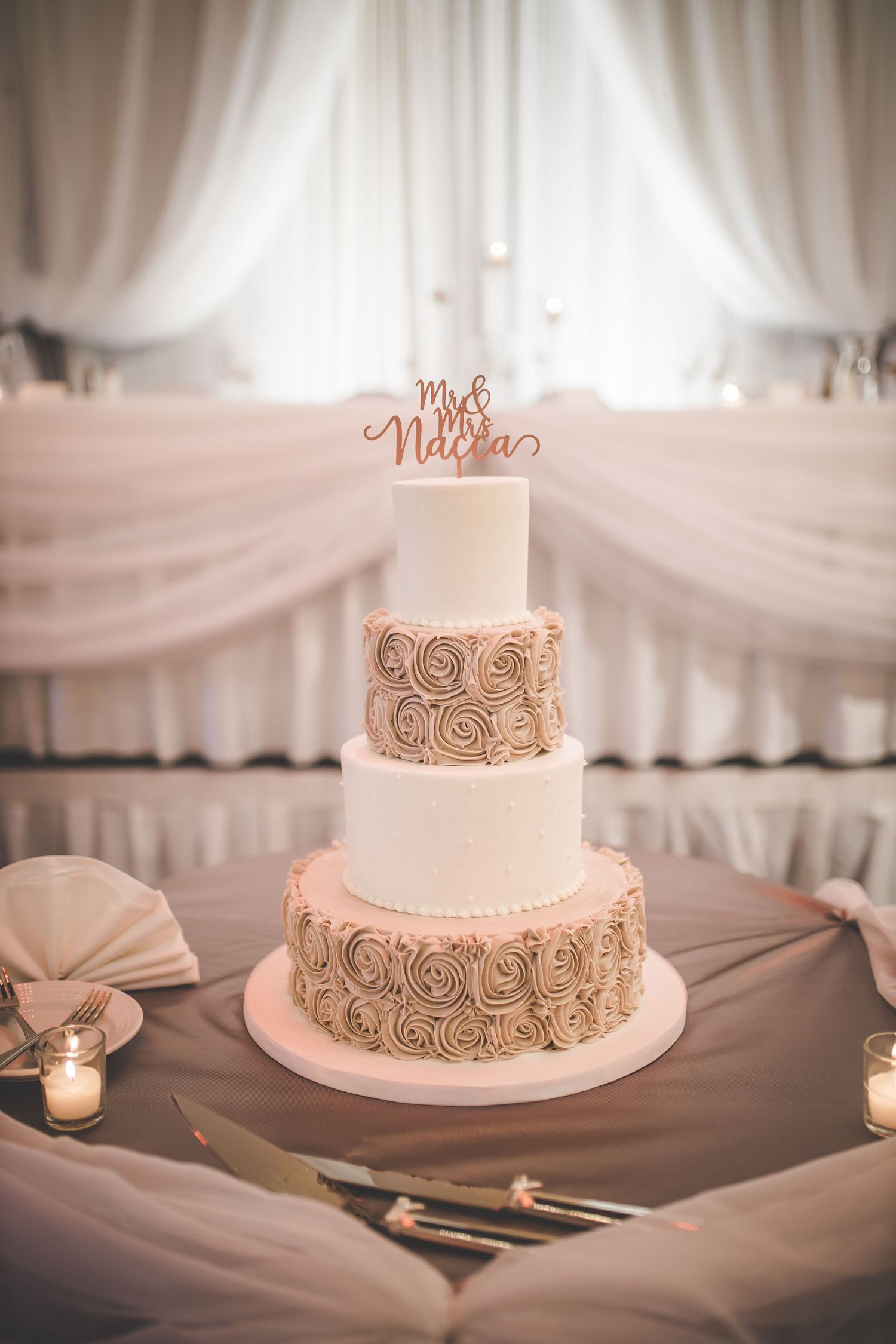 An elegant two-tier white wedding cake with rose gold accents displayed on a draped table.