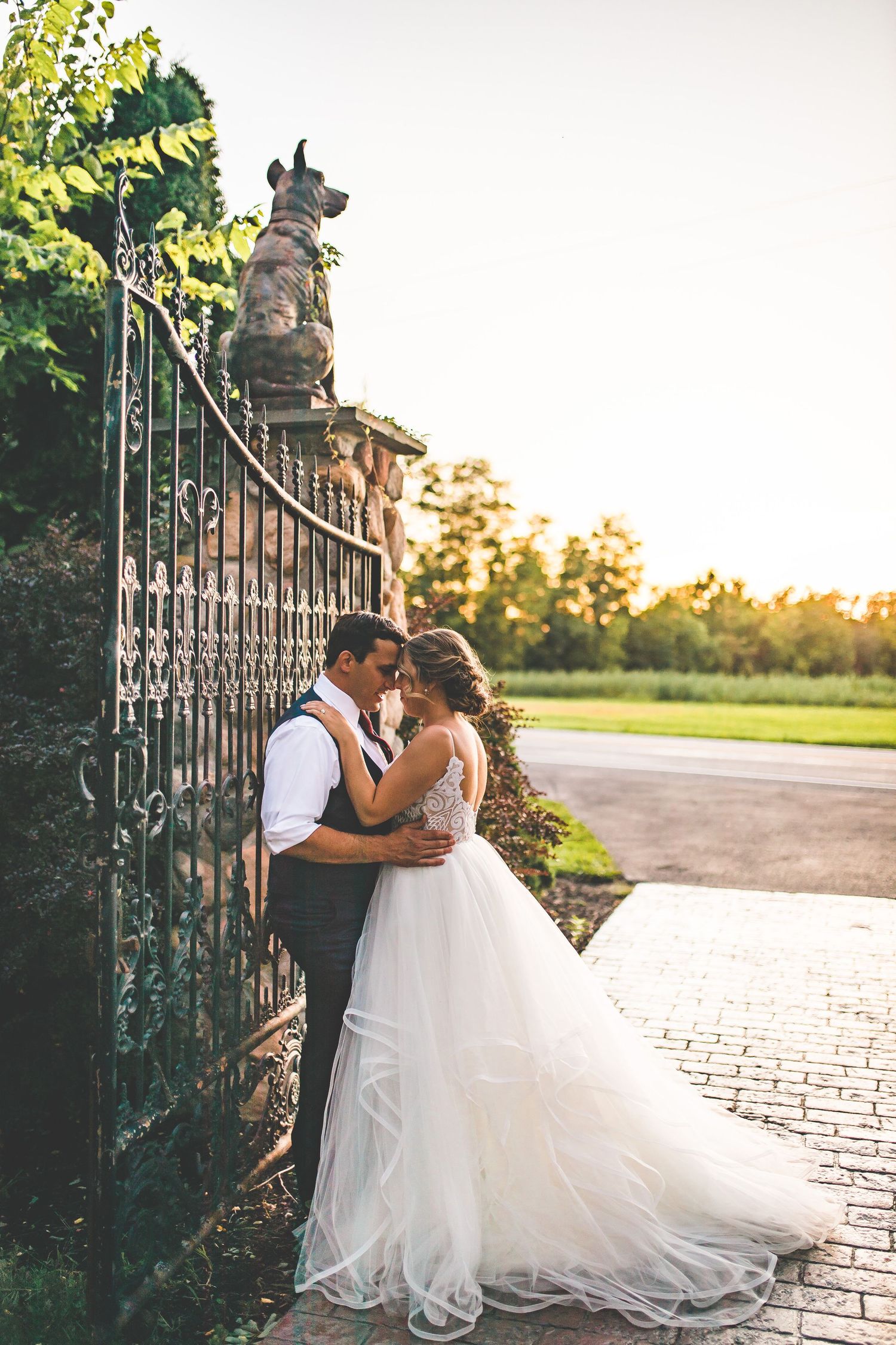 Romantic wedding couple embracing by an ornate iron gate at sunset.