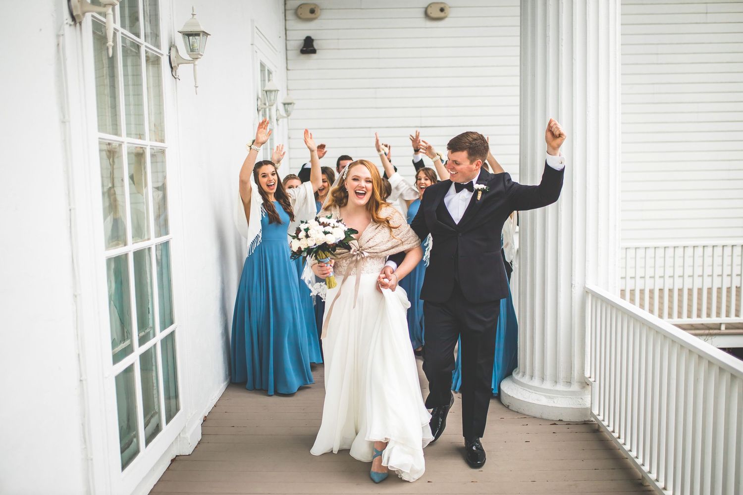 Wedding party celebrates joyfully on a white columned porch.