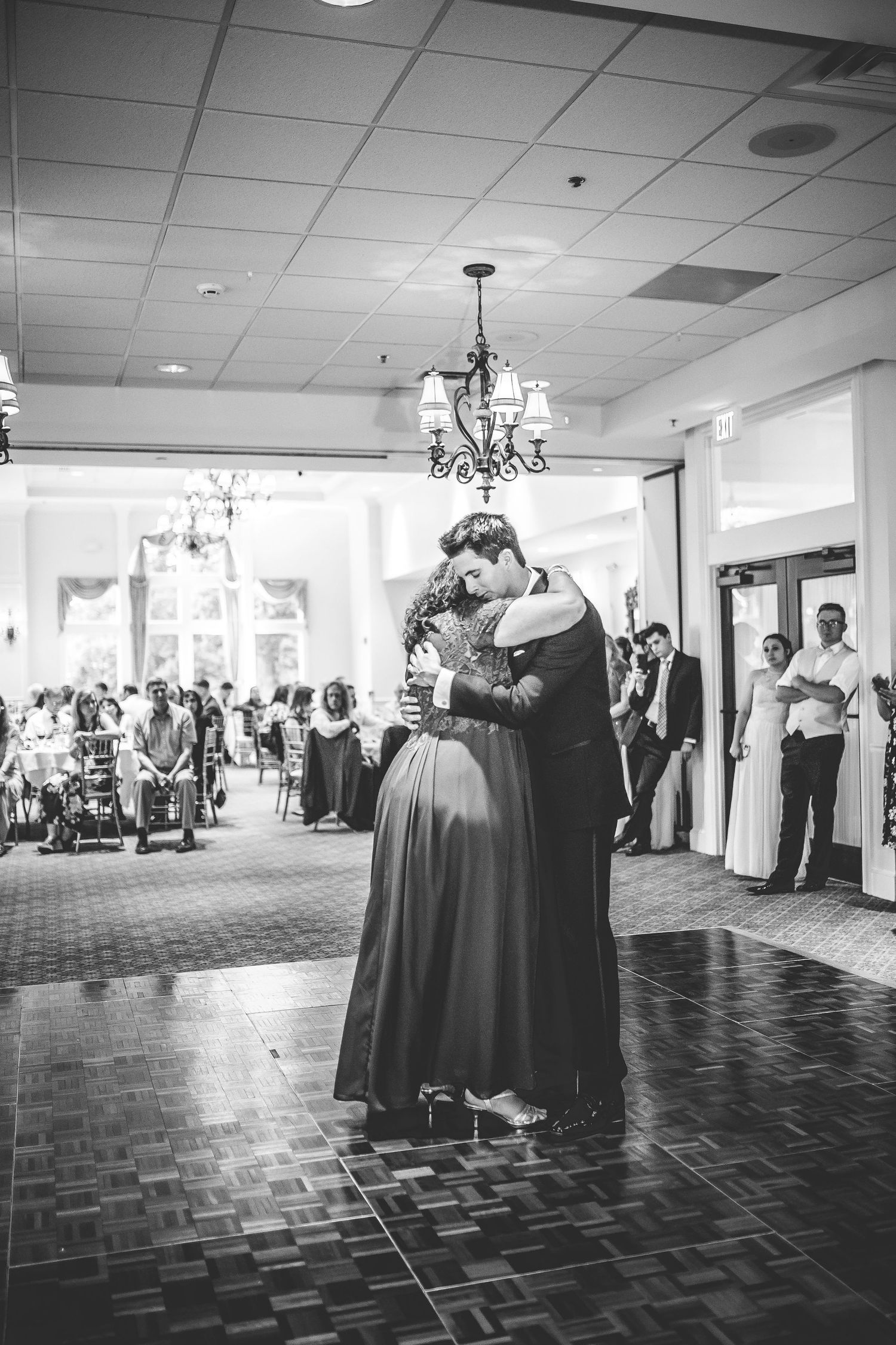 Black and white photo of a wedding dance in an elegant ballroom with chandeliers and guests looking on.