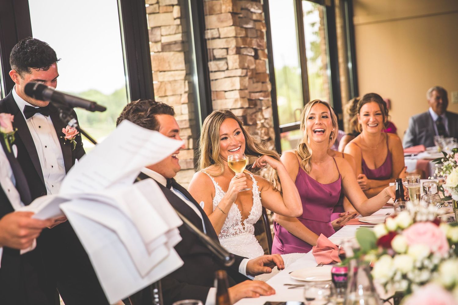 Wedding guests laugh and smile during speeches at an elegant reception with floral centerpieces.