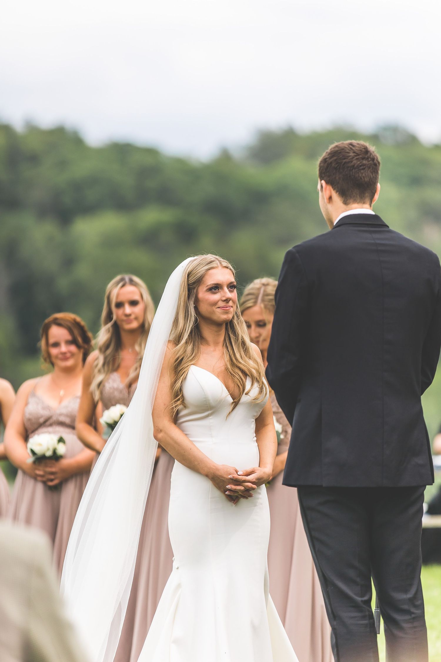 Bridesmaids in blush dresses stand beside bride in white gown during outdoor ceremony.