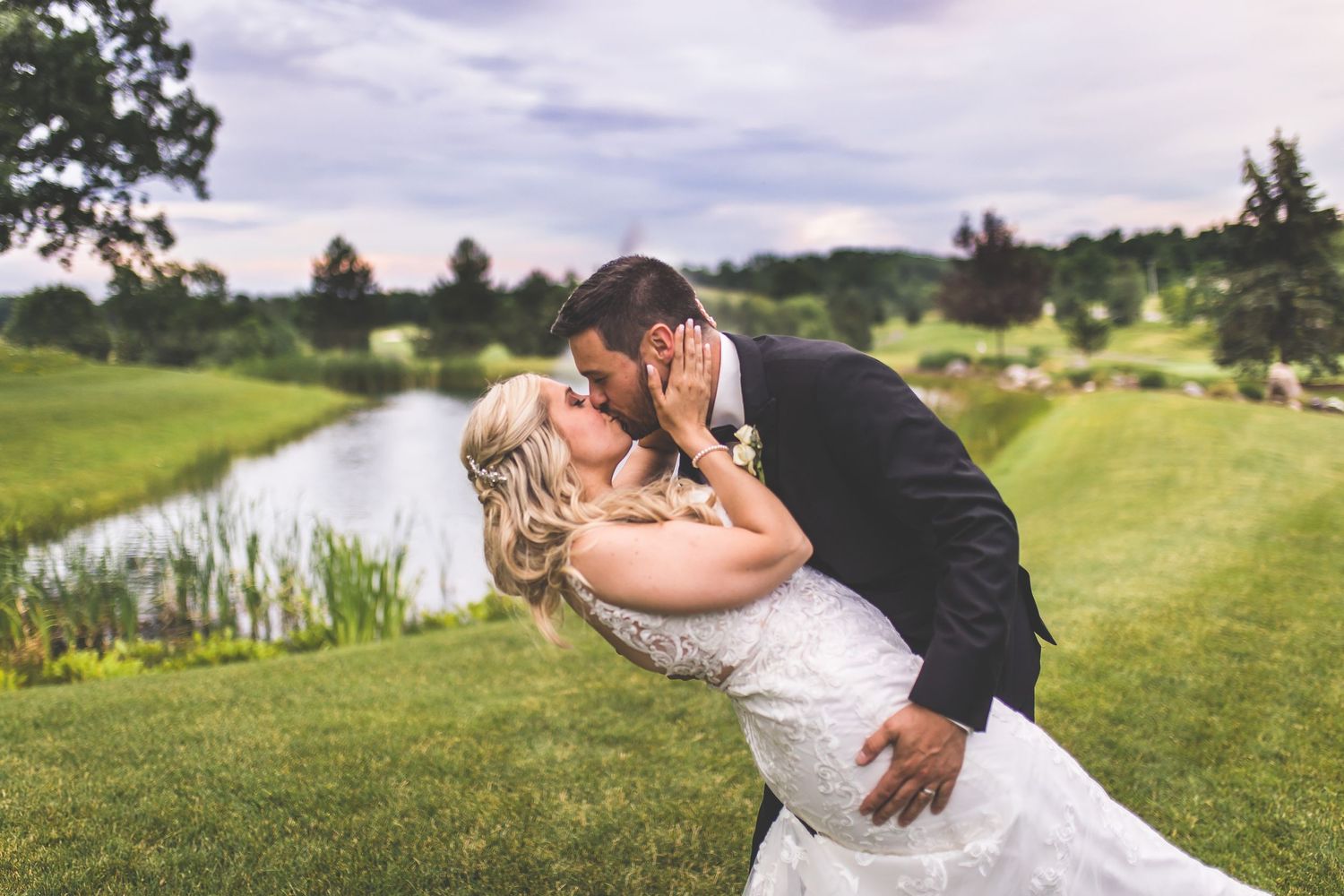 A romantic wedding couple embraces on a scenic golf course with rolling hills and a lake in the background.