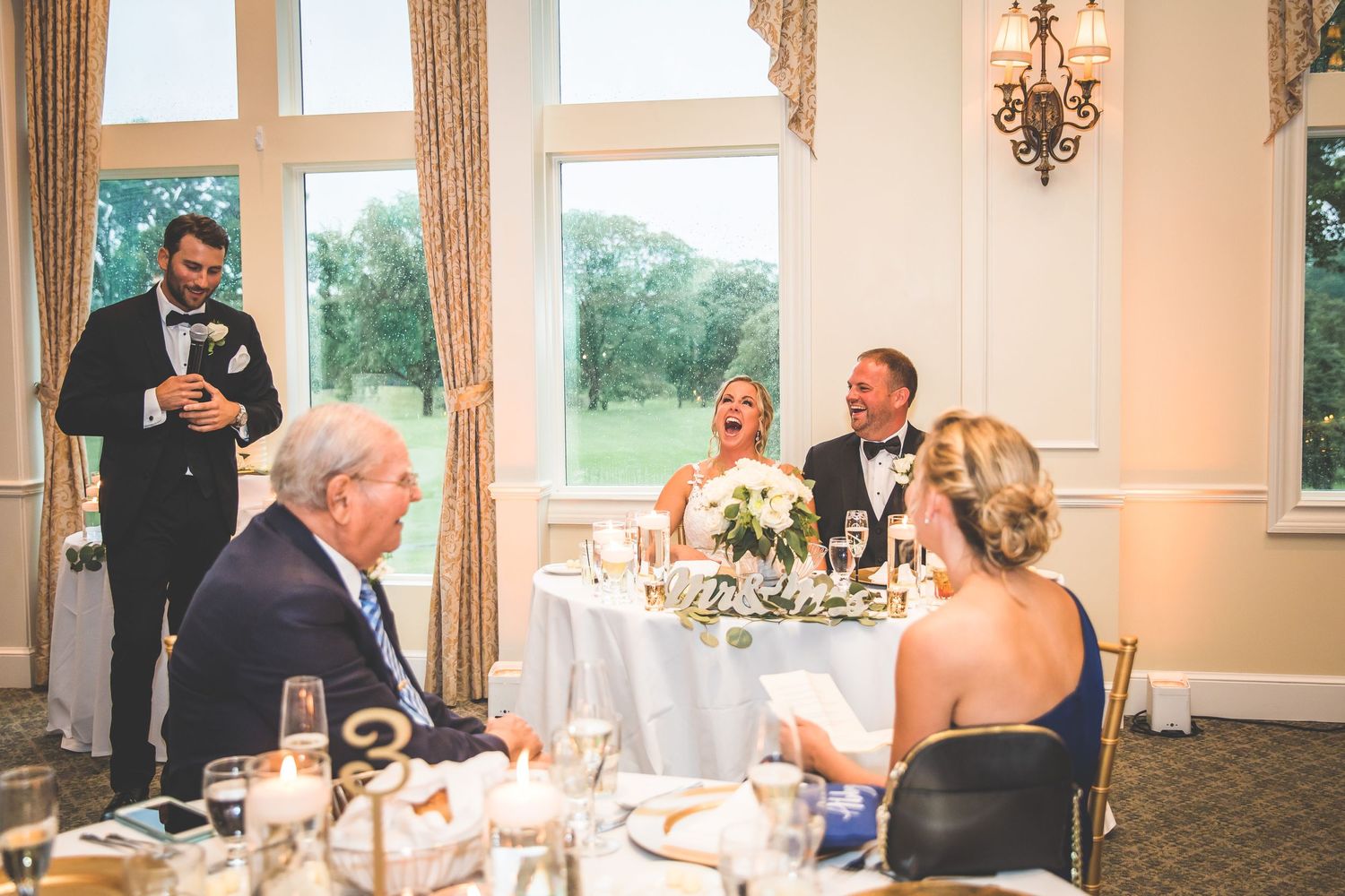 Wedding reception toast and speeches during an elegant dinner with large windows and natural light.