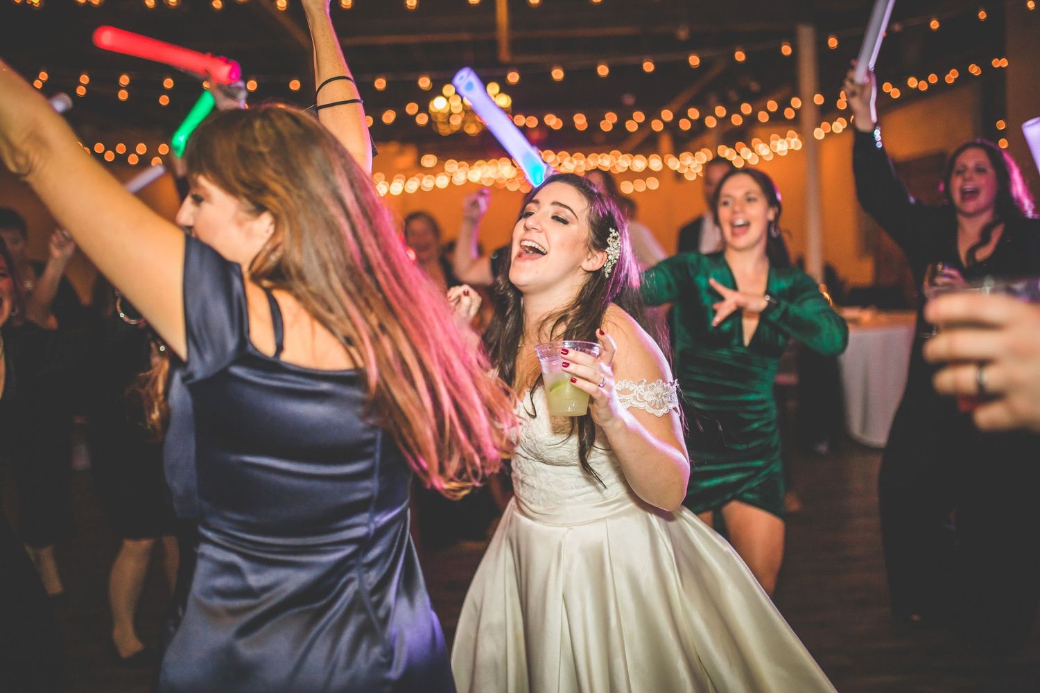 Lively dance party at wedding reception with string lights overhead and guests dancing.
