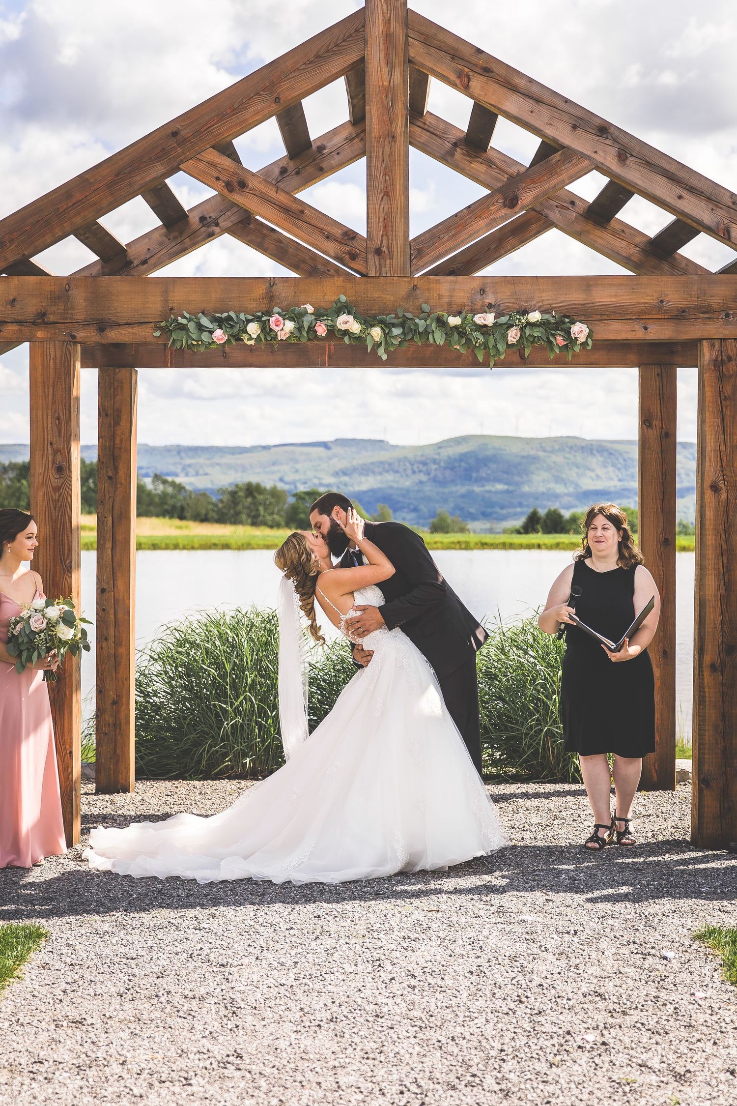 A rustic wooden pergola decorated with floral garlands frames a wedding ceremony against mountain views.