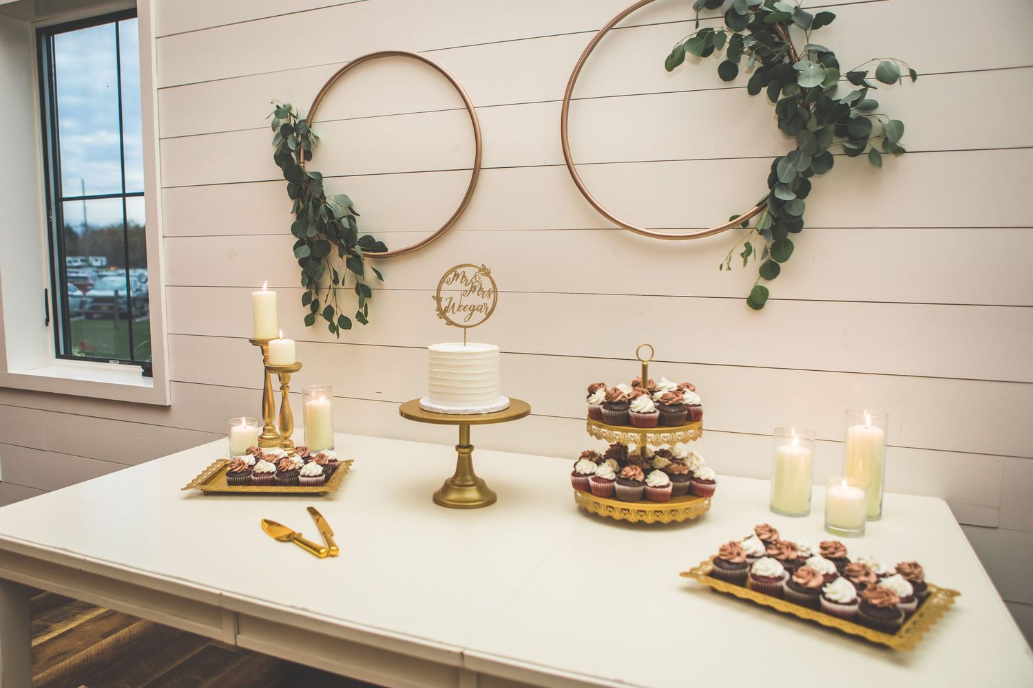 Wedding dessert display with gold accents and greenery on white tables against shiplap walls.