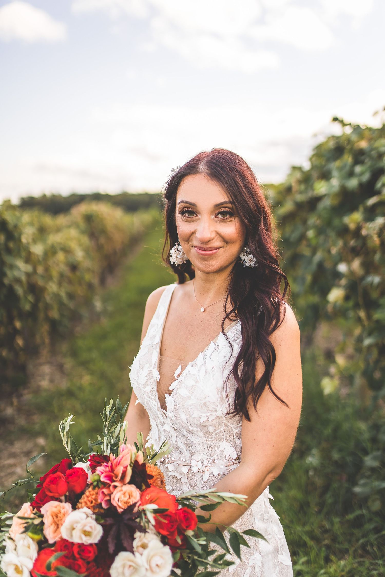 Bride with red and pink bouquet smiles during vineyard wedding photoshoot.