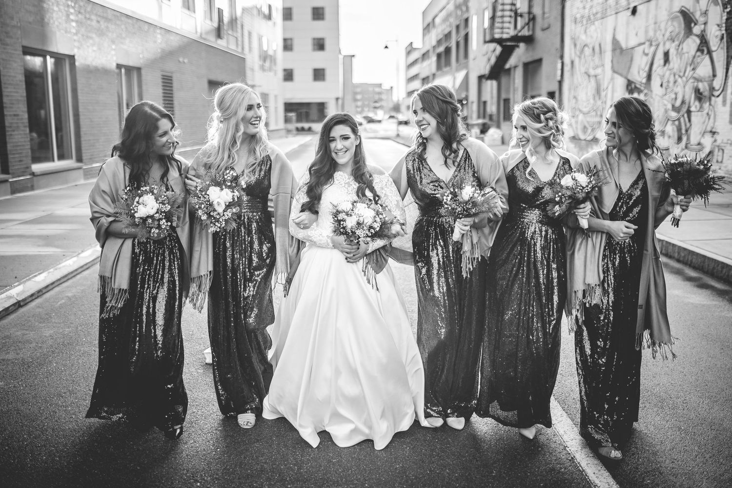 Wedding party bridesmaids in dark dresses laugh together on city street.