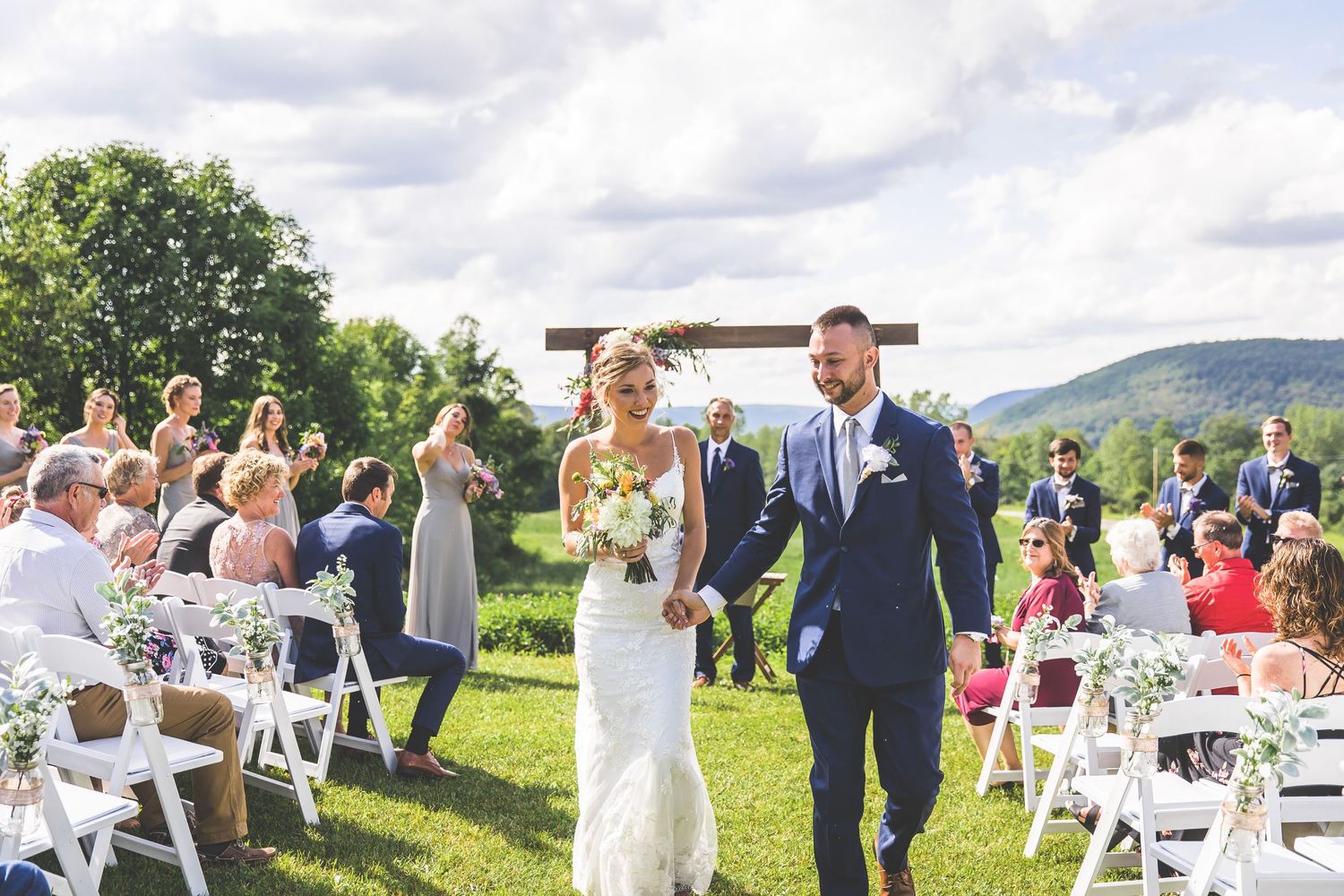 Wide angle view of wedding guests and ceremony site on rolling hills under cloudy sky.